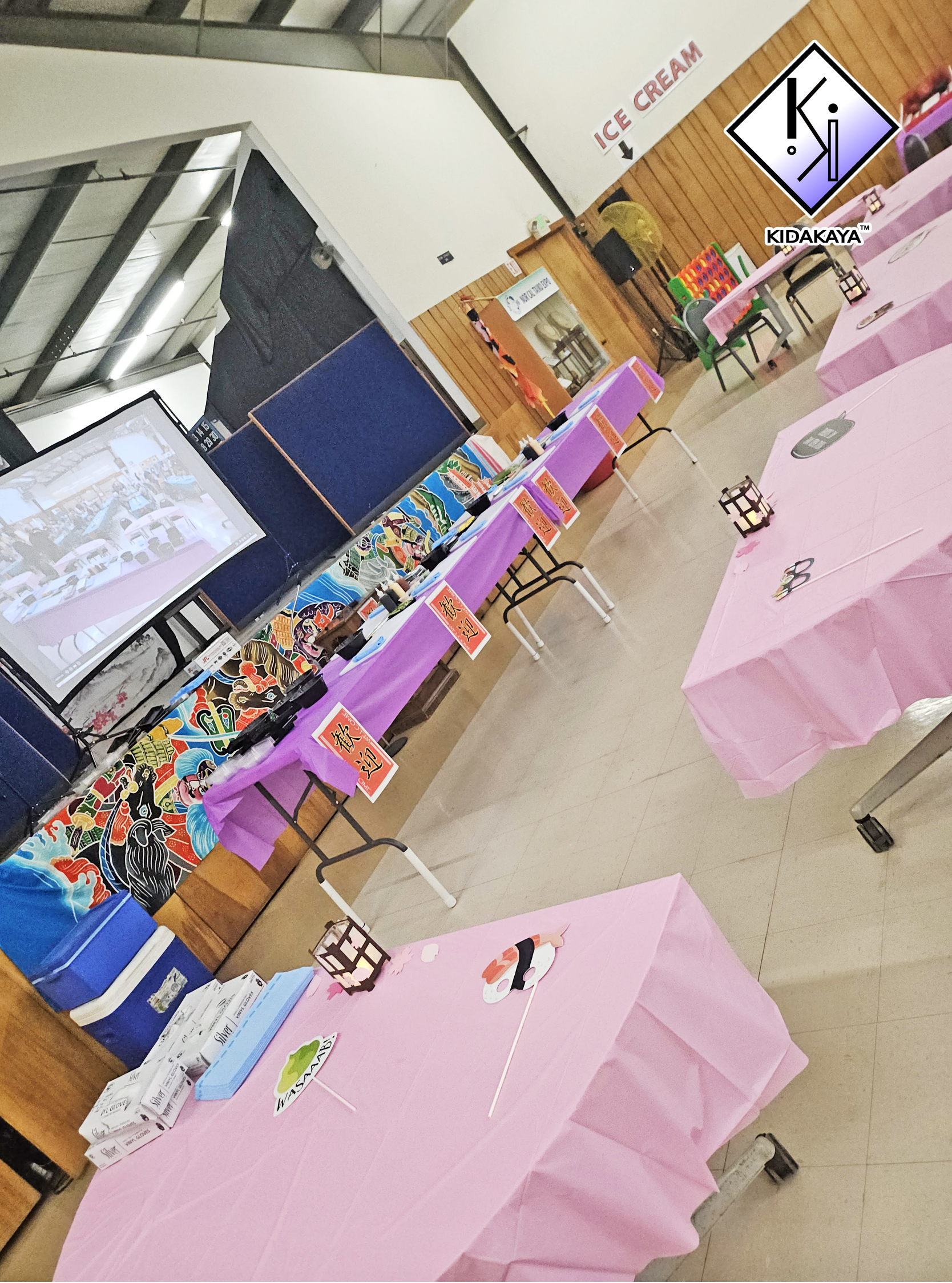 A room decorated for a celebration with tables covered in pink tablecloths, witchcraft-themed decorations, and signs with names like "WASABI" and various labels. There is a projection screen showing an image of the same room, and a colorful game on a