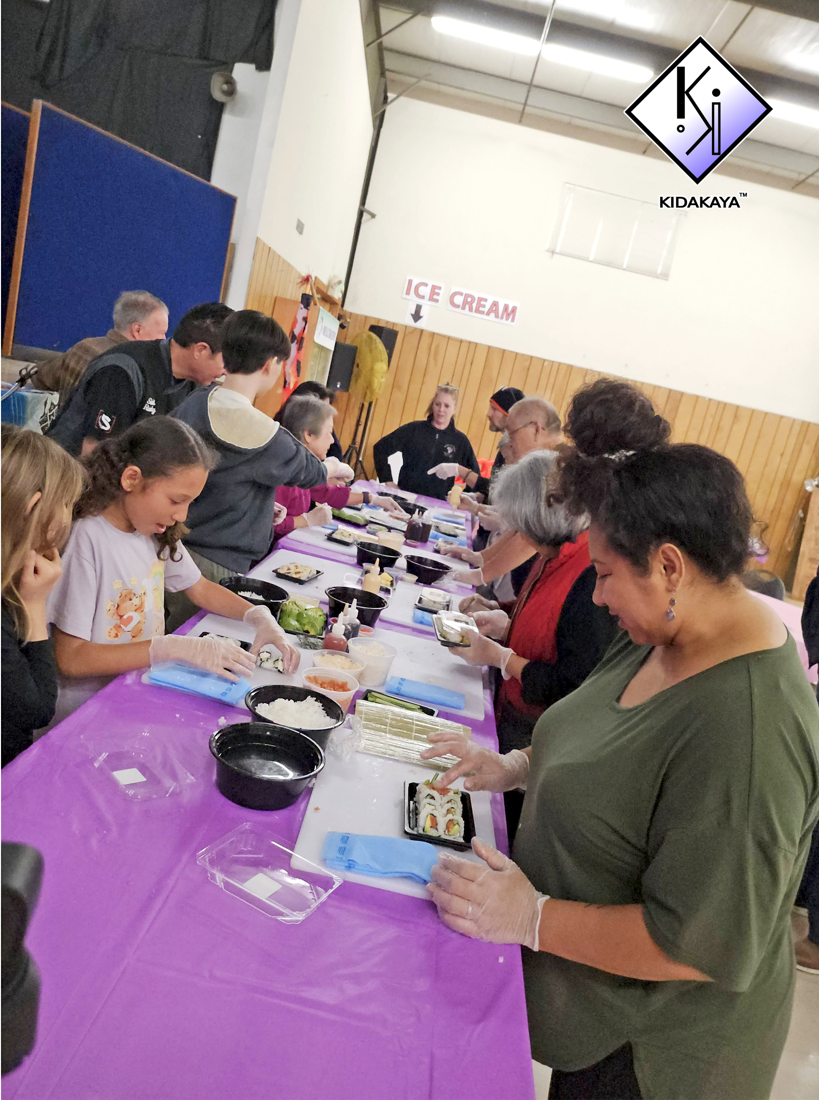 People gathered around a table making sushi at a sushi-making workshop in a community center with wooden-paneled walls, a sign labeled 'ICE CREAM', and a purple tablecloth on the table.