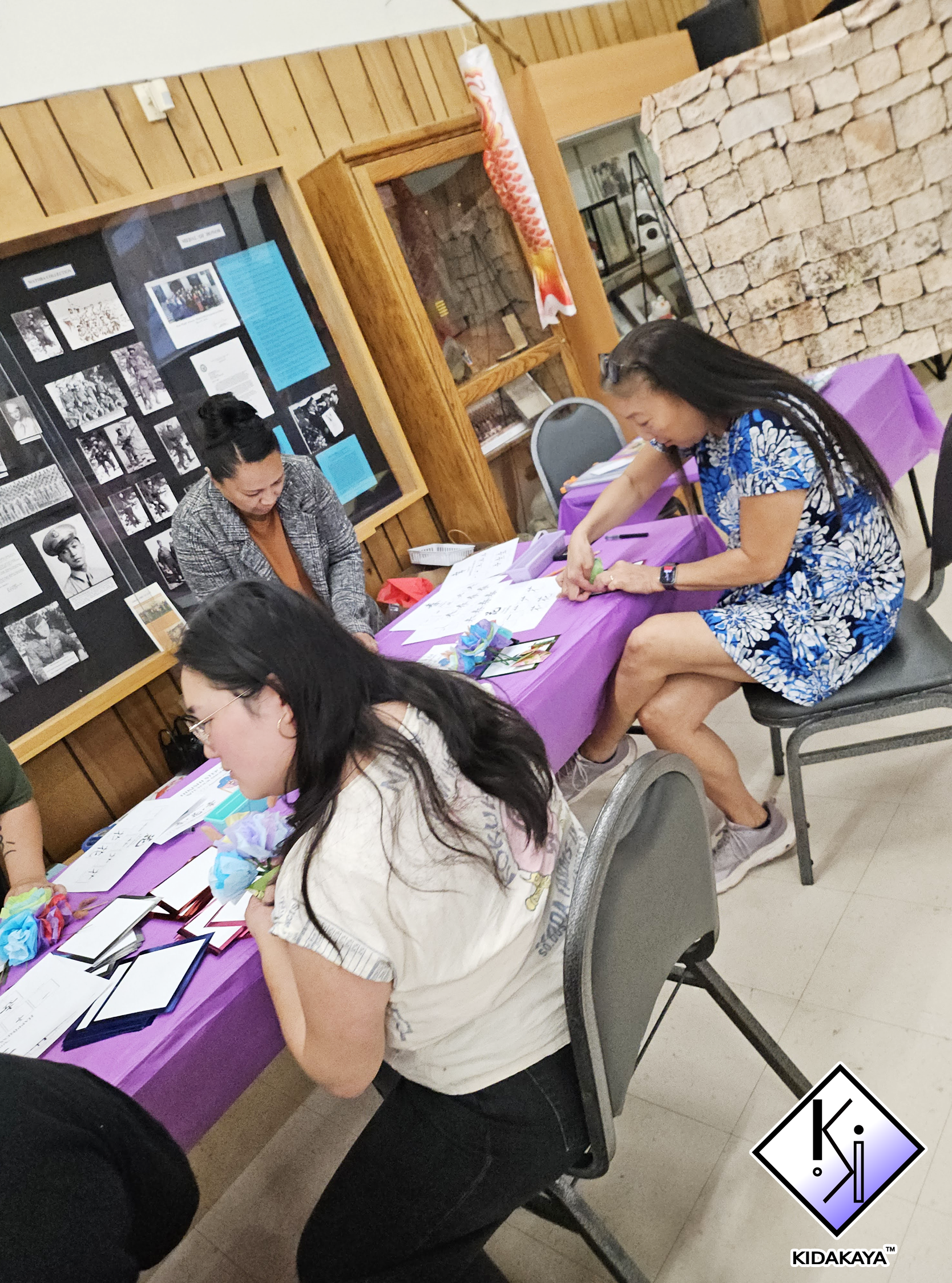 A group of women sitting and standing around a table covered with a purple tablecloth, engaging in arts and crafts activities, with a display board of black and white photographs and documents on a wooden wall behind them.