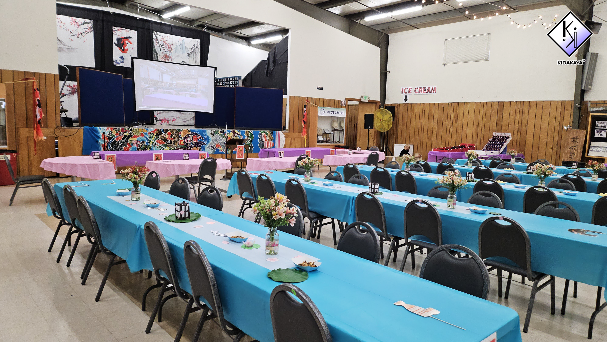 A decorated banquet hall with long tables covered in blue tablecloths, adorned with flower arrangements and food bowls. The background features a stage with traditional Asian decorations, posters, and a projection screen. The hall has wooden paneling