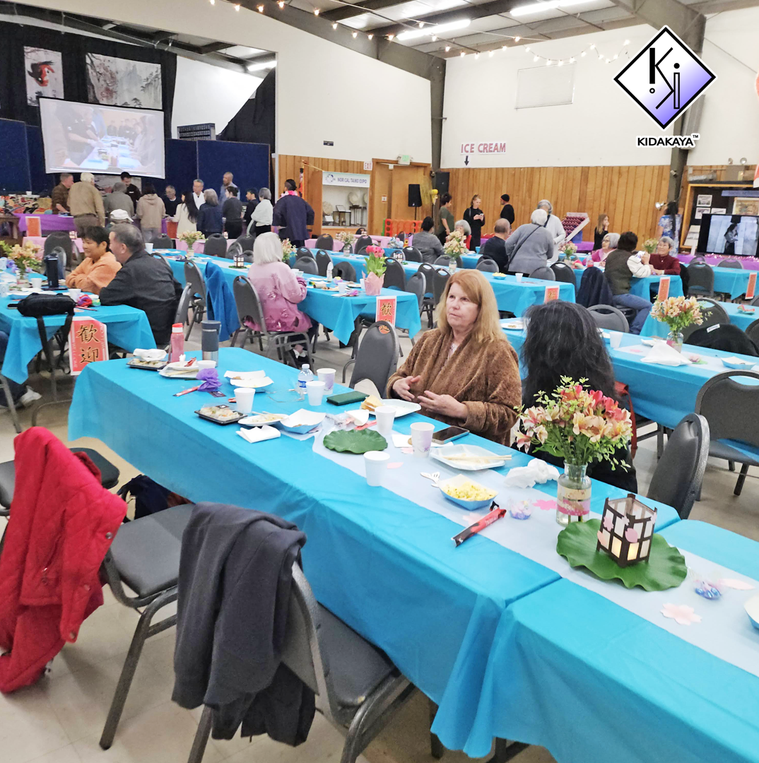 People dining at a banquet with blue tablecloths, floral centerpieces, and a decorated indoor setting, featuring a large projected screen and signs for ice cream and food.