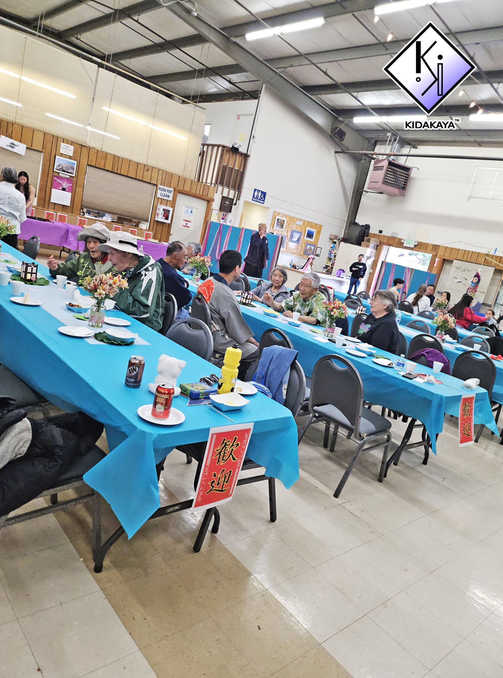 Indoor event with people seated at tables covered with blue tablecloths, decorated with flower centerpieces. Signage and artwork on the walls, with an entrance or exit in the background. Some attendees are engaged in conversations, and a few are look