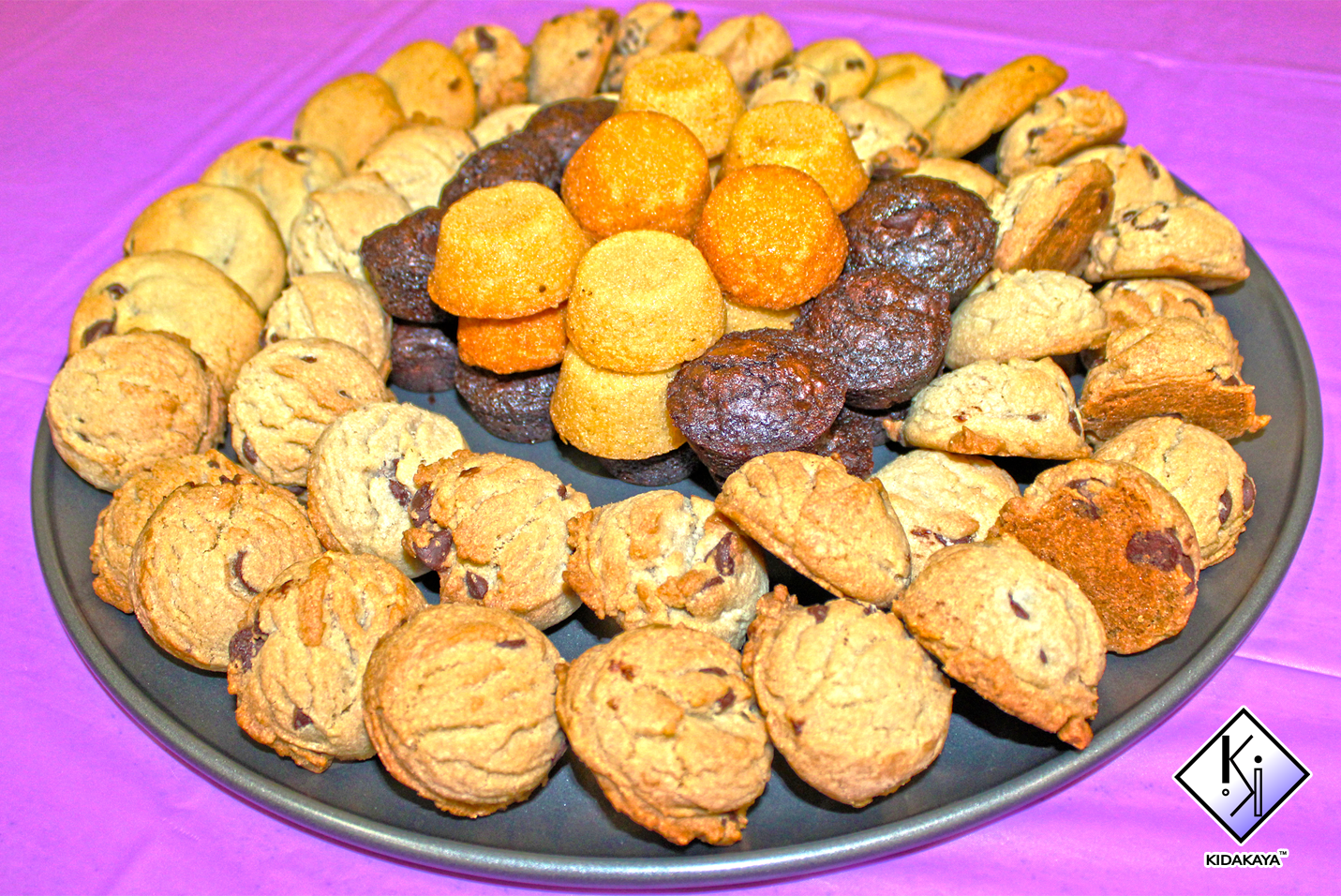 A large black platter filled with an assortment of cookies, including chocolate chip, chocolate, and sandwich cookies, arranged in a circular pattern on a purple tablecloth.