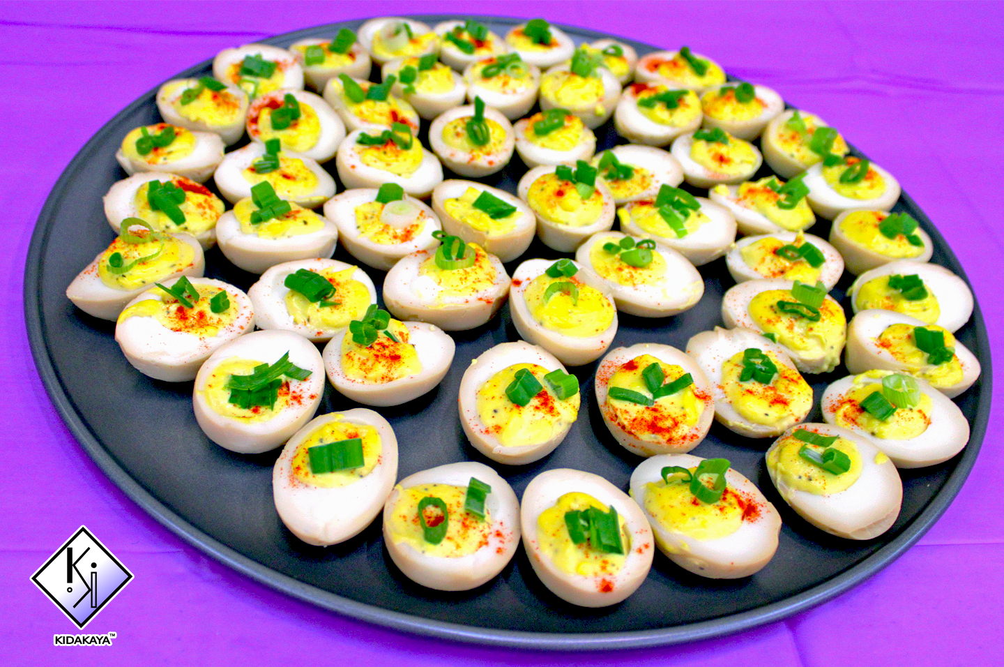 A black tray filled with halved boiled eggs topped with chopped green onions and paprika on a purple surface.