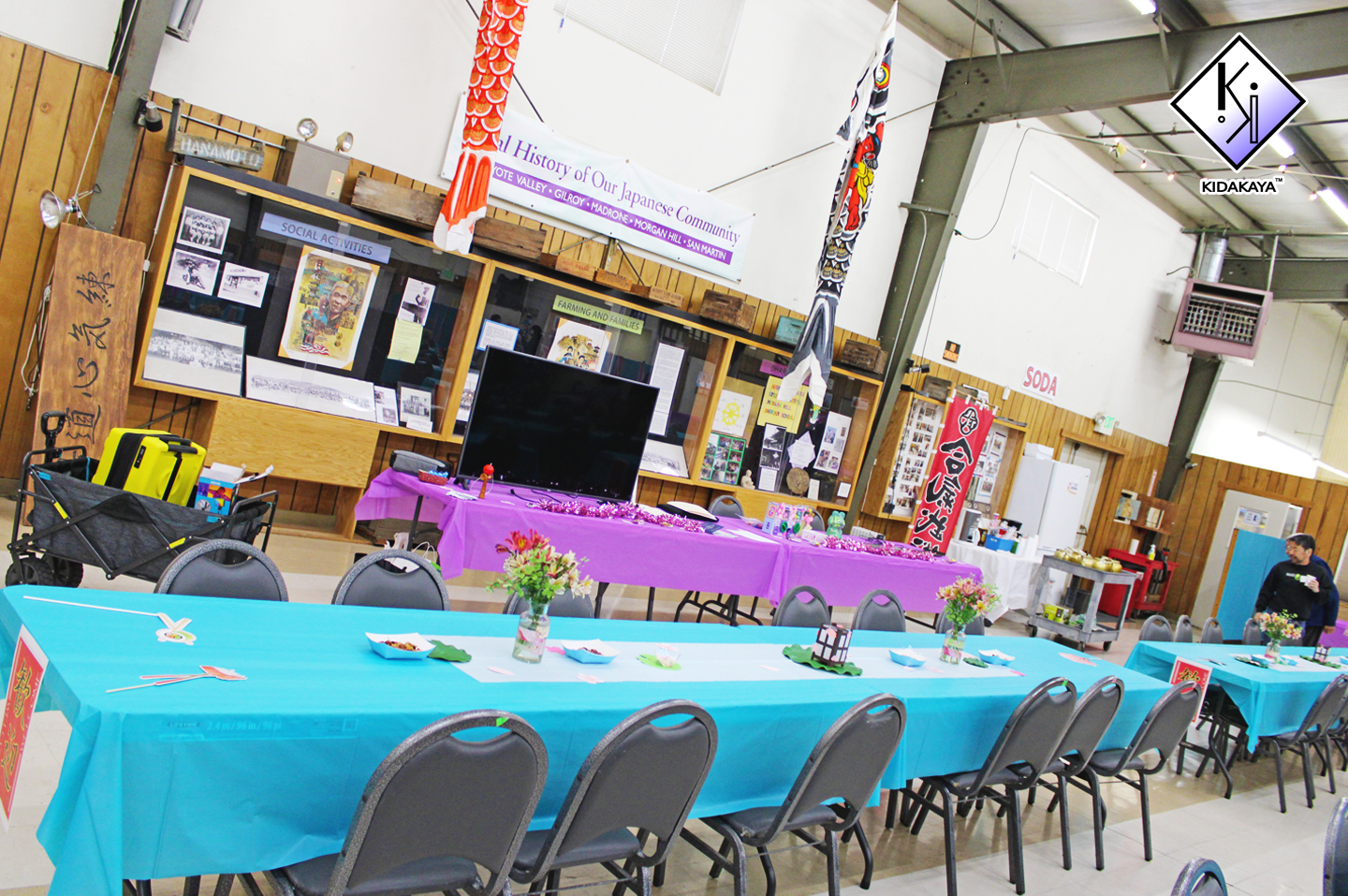 Indoor event setup with colorful tables, chairs, and decorations, including banners and display cases on the back wall. There are flowers on the tables and a large TV in the center.