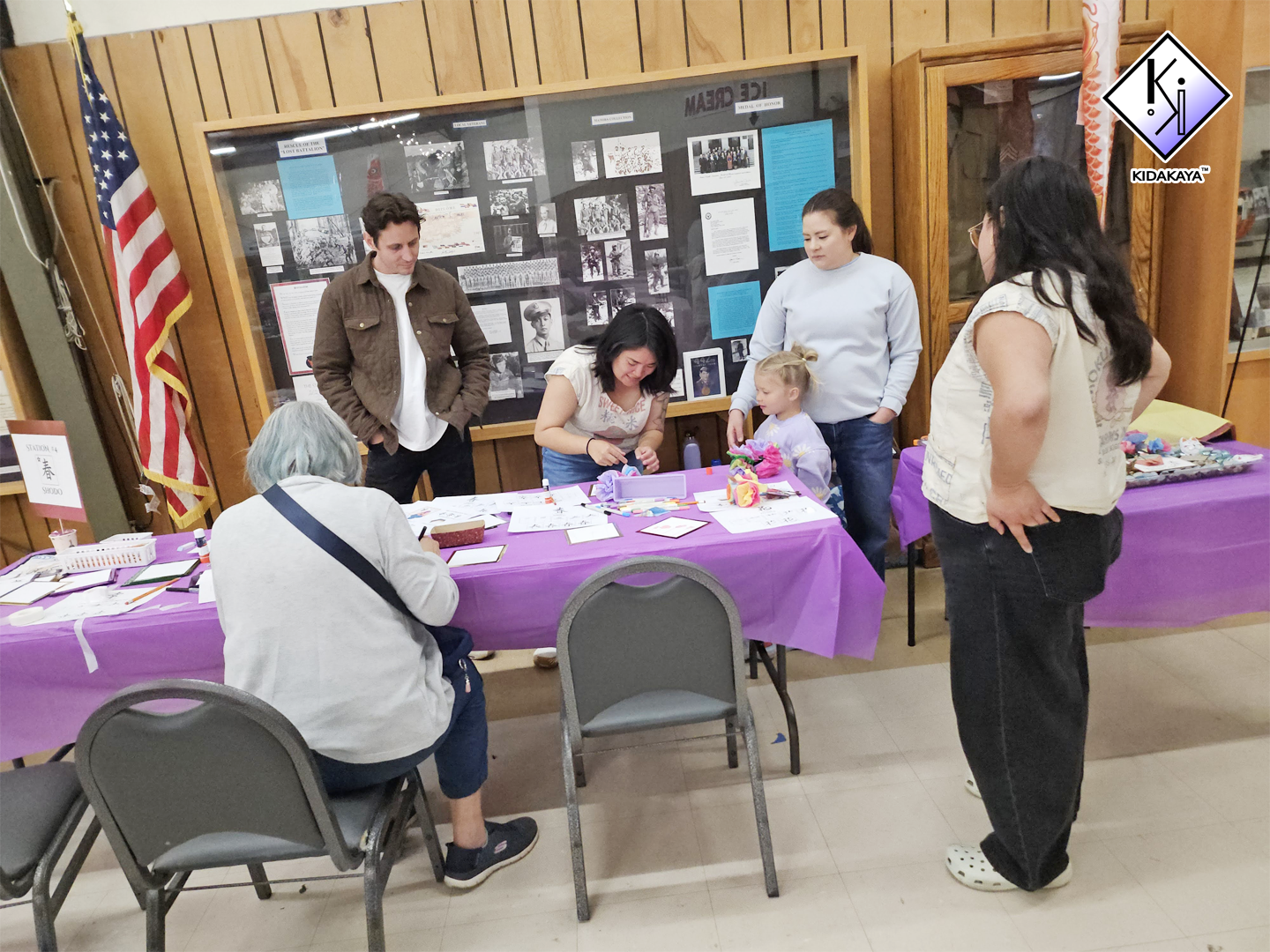 People gathered around a table with crafts and papers, inside a room with wood-paneled walls, an American flag, and a display board behind them.