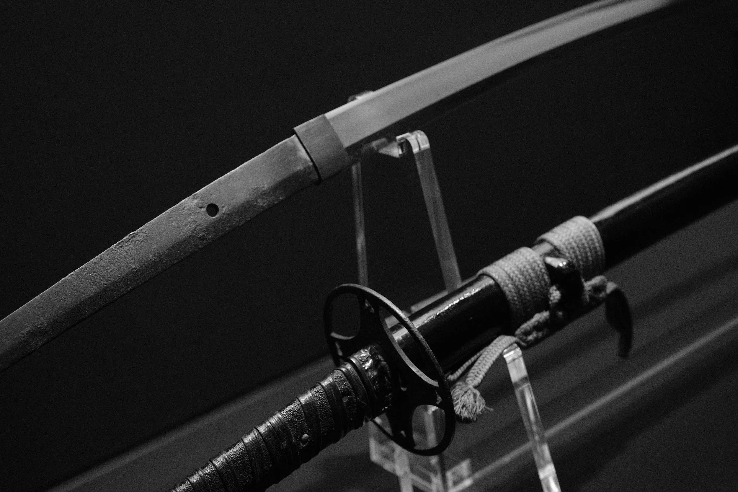 Close-up black and white image of a katana mounted on a display rack against a dark background.