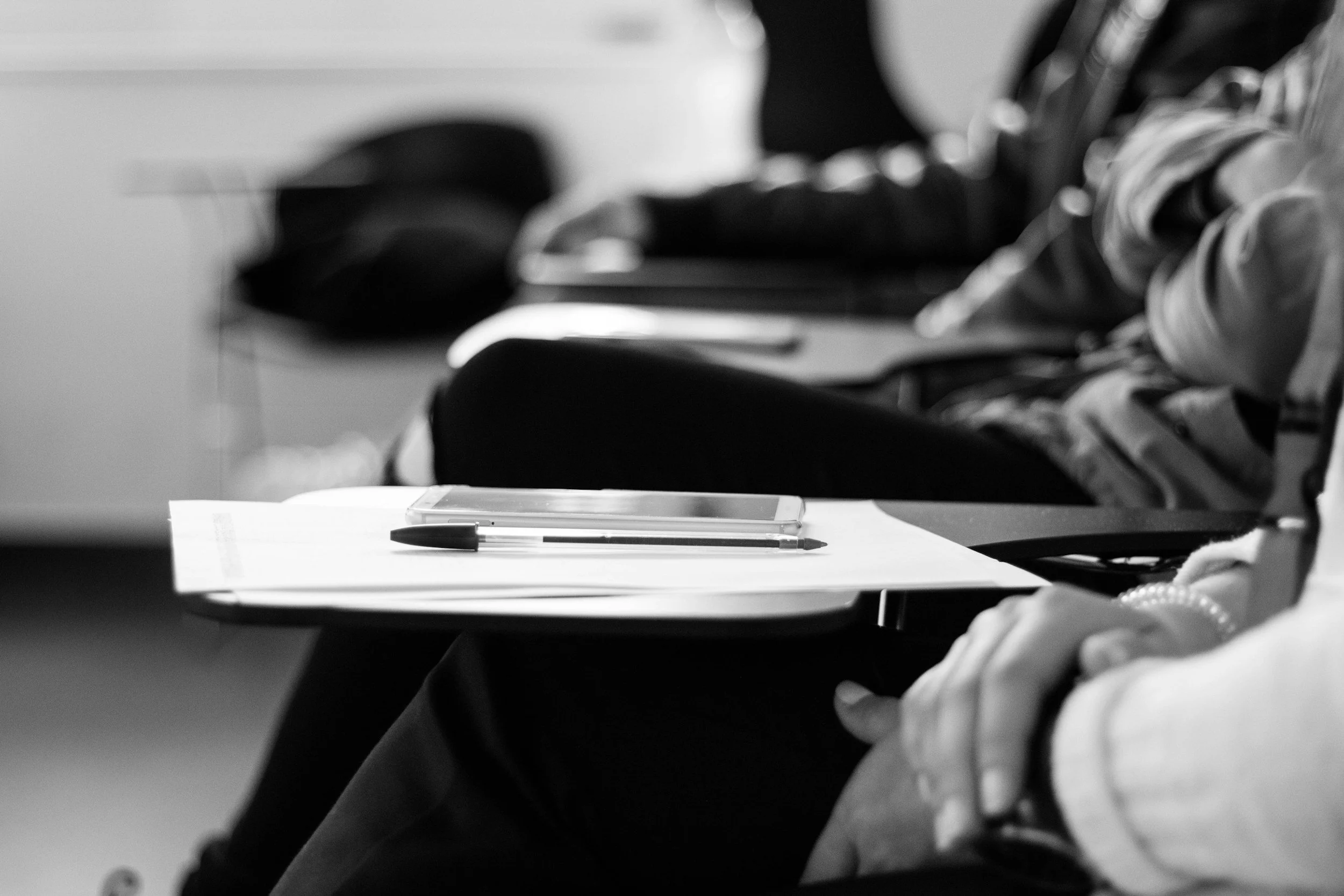A person sitting at a table with a pen and paper, with other people in the background, blurred.