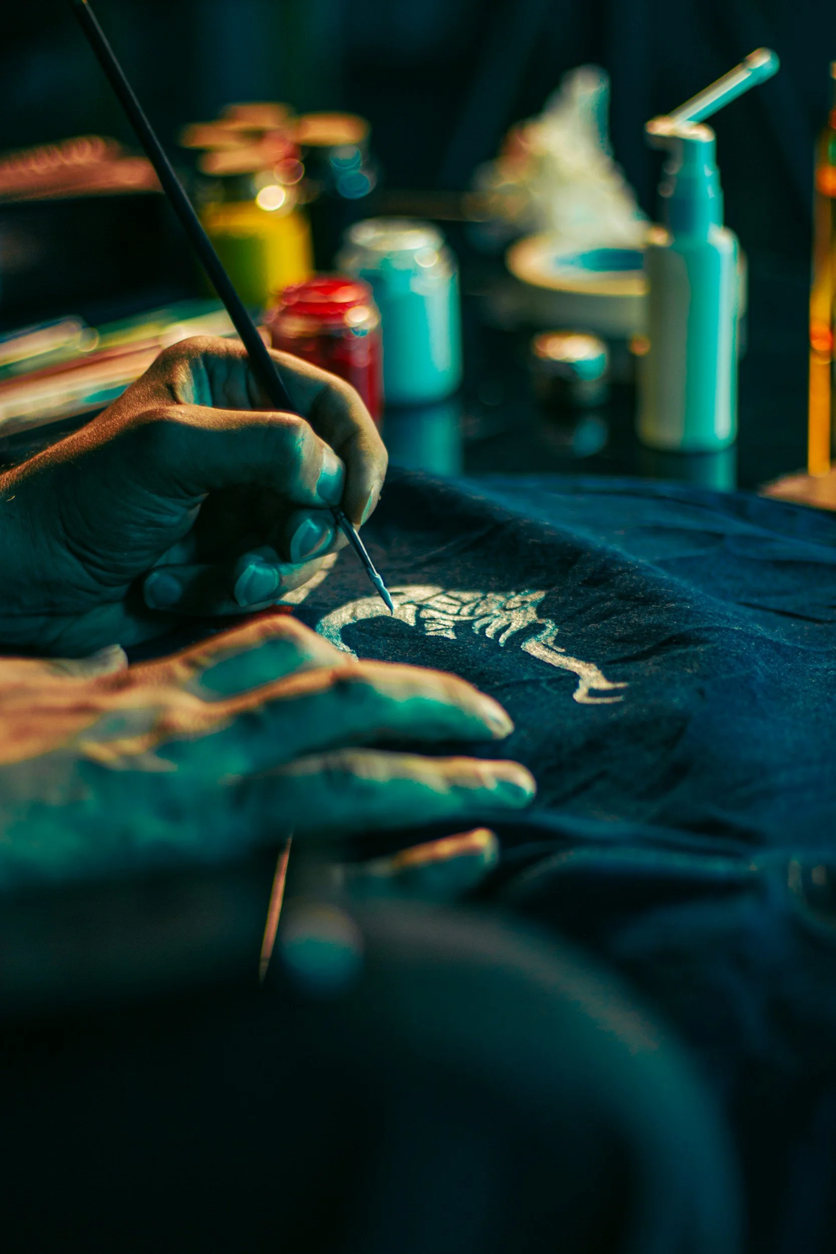 Close-up of an artist painting on canvas with a fine brush, in a dimly lit studio, with cans of paint in the background.