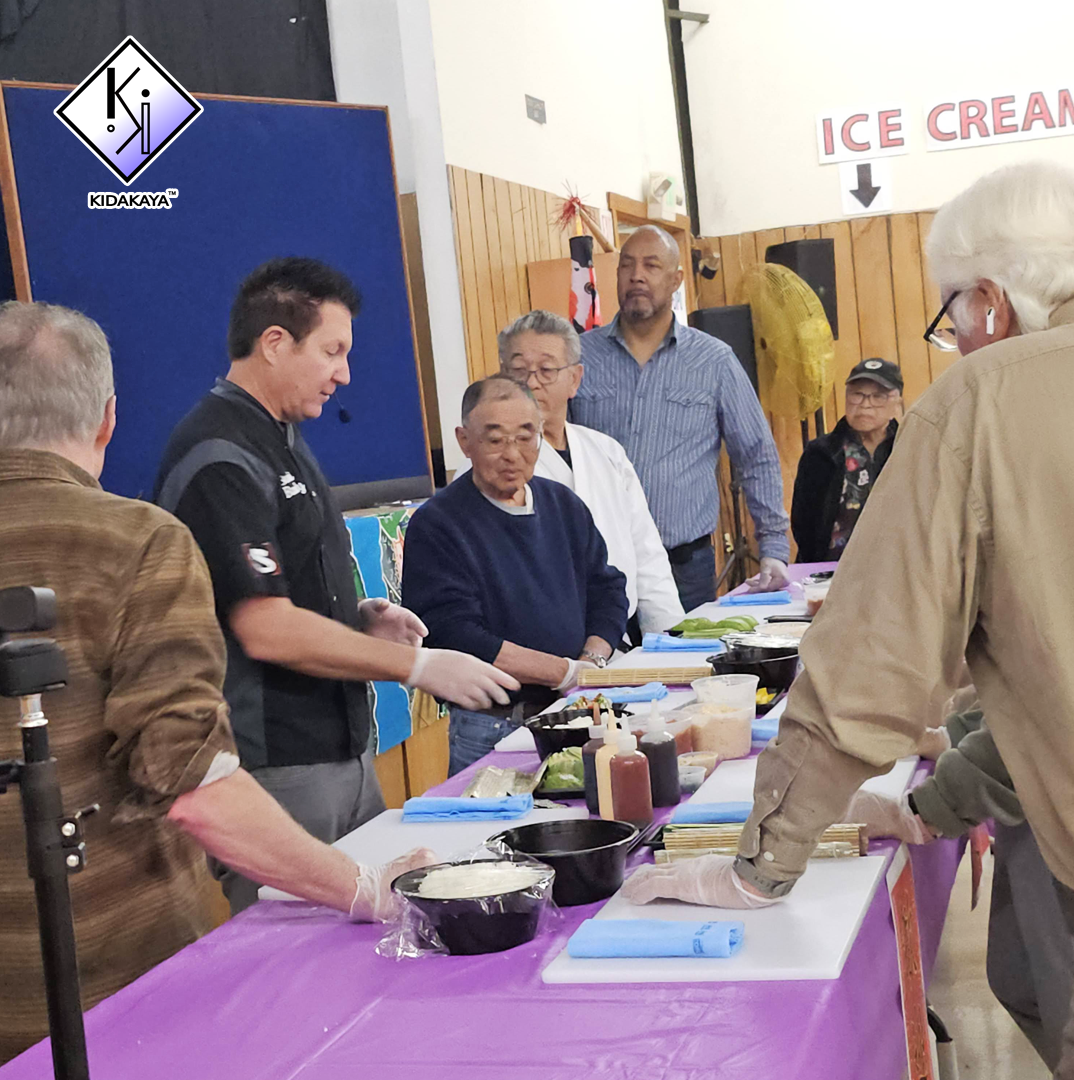 Group of people gathered around a table with food ingredients, some wearing gloves, in a community center or event space, with a sign that says 'Ice Cream' in the background.