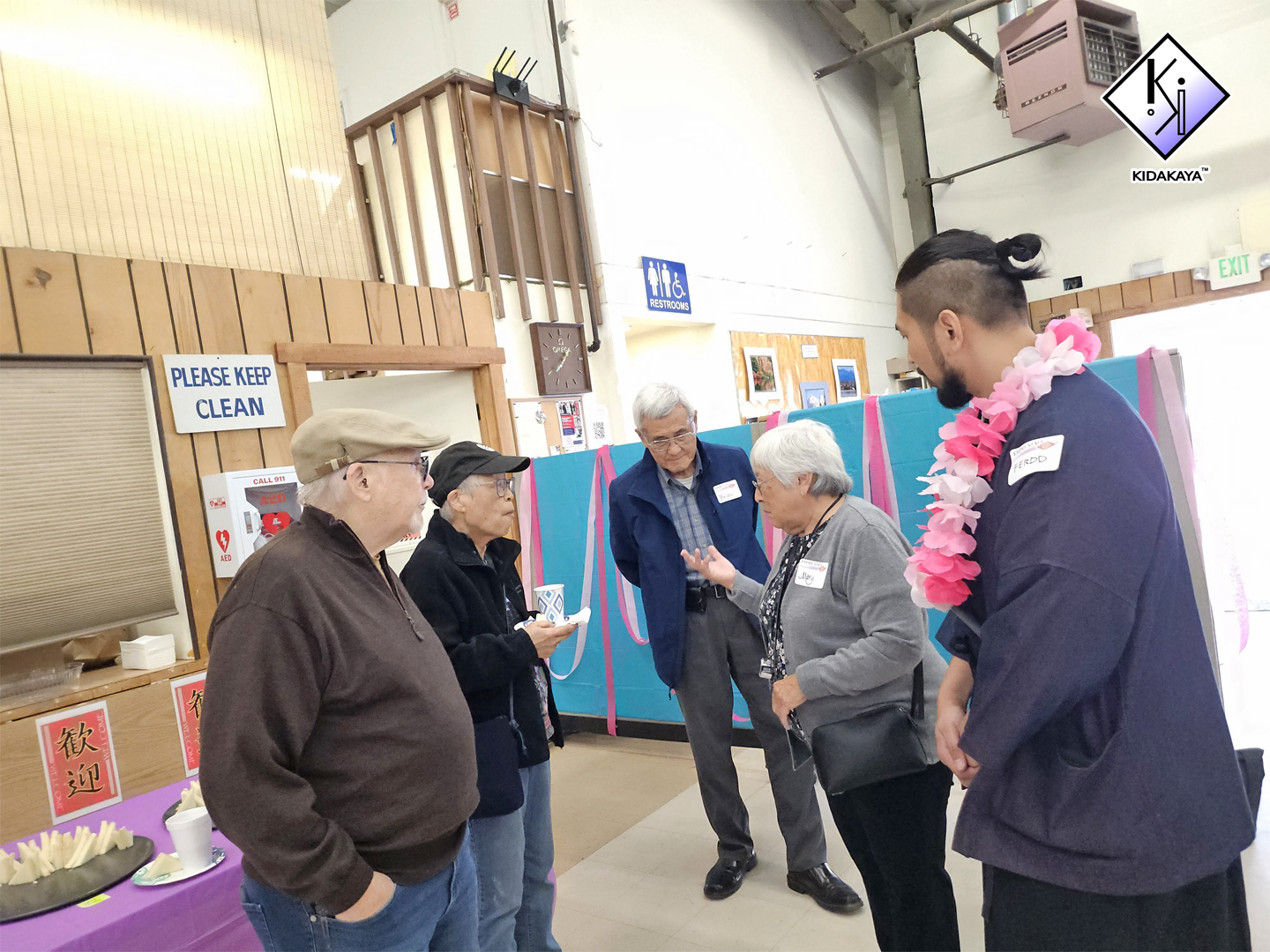 Group of five people standing and talking indoors, decorated with pink and blue streamers, near a table with food and cups, in what appears to be a celebration or gathering.