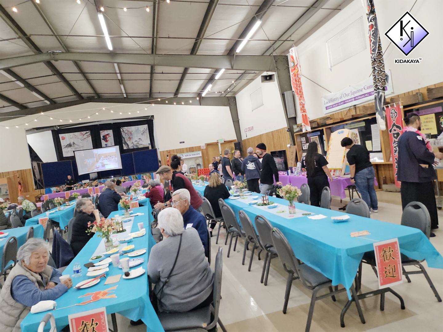 A large indoor gathering with tables covered in blue and purple tablecloths, decorated with flowers and paper lanterns, and people sitting and standing around, eating and socializing in a spacious hall with a high ceiling and string lights.