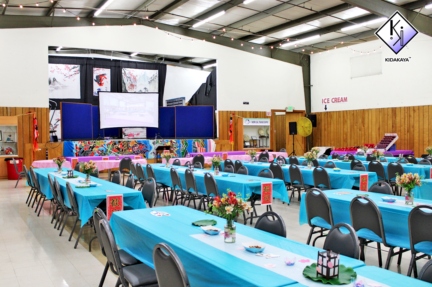 Banquet hall decorated with blue and pink tablecloths, floral centerpieces, and Japanese-themed artwork on the walls, with a stage in the background and an ice cream sign on the wall.