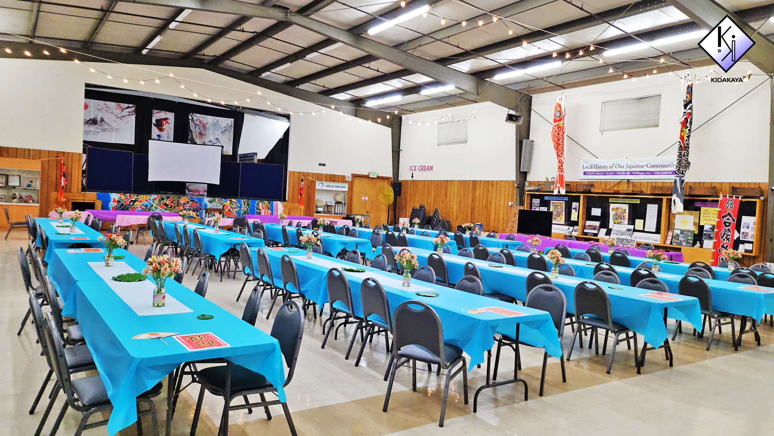 A spacious event hall decorated with blue tablecloths, floral centerpieces, and festive decorations for a Japanese community gathering.