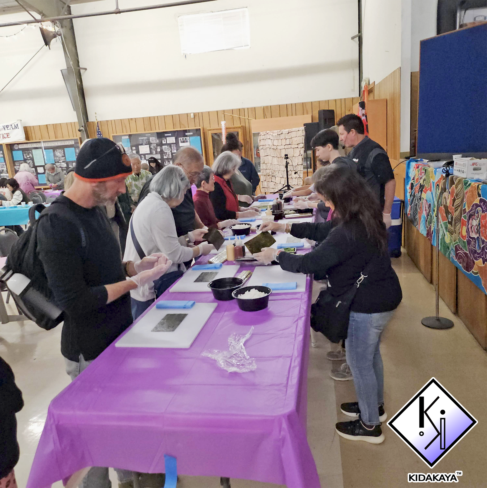 People gathered around a long purple table in an indoor community hall, participating in a hands-on activity, with display boards and decorated walls in the background.