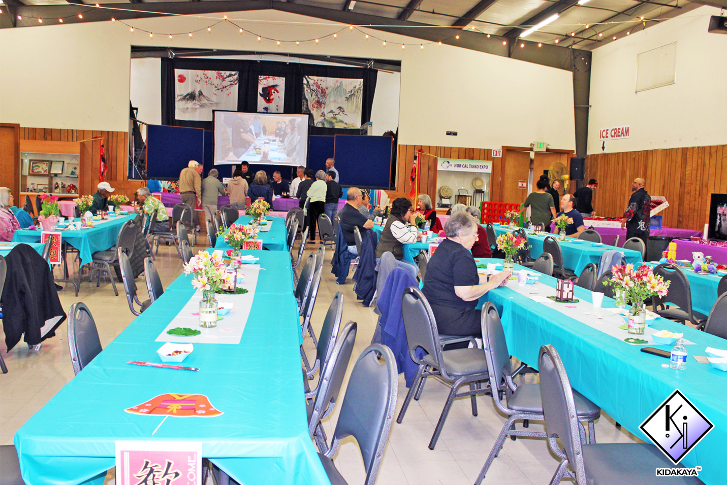 Chuukese community gathering with seated guests at colorful tables, some standing near a stage with large projection screen, in a decorated indoor event space with string lights and artwork on the back wall.