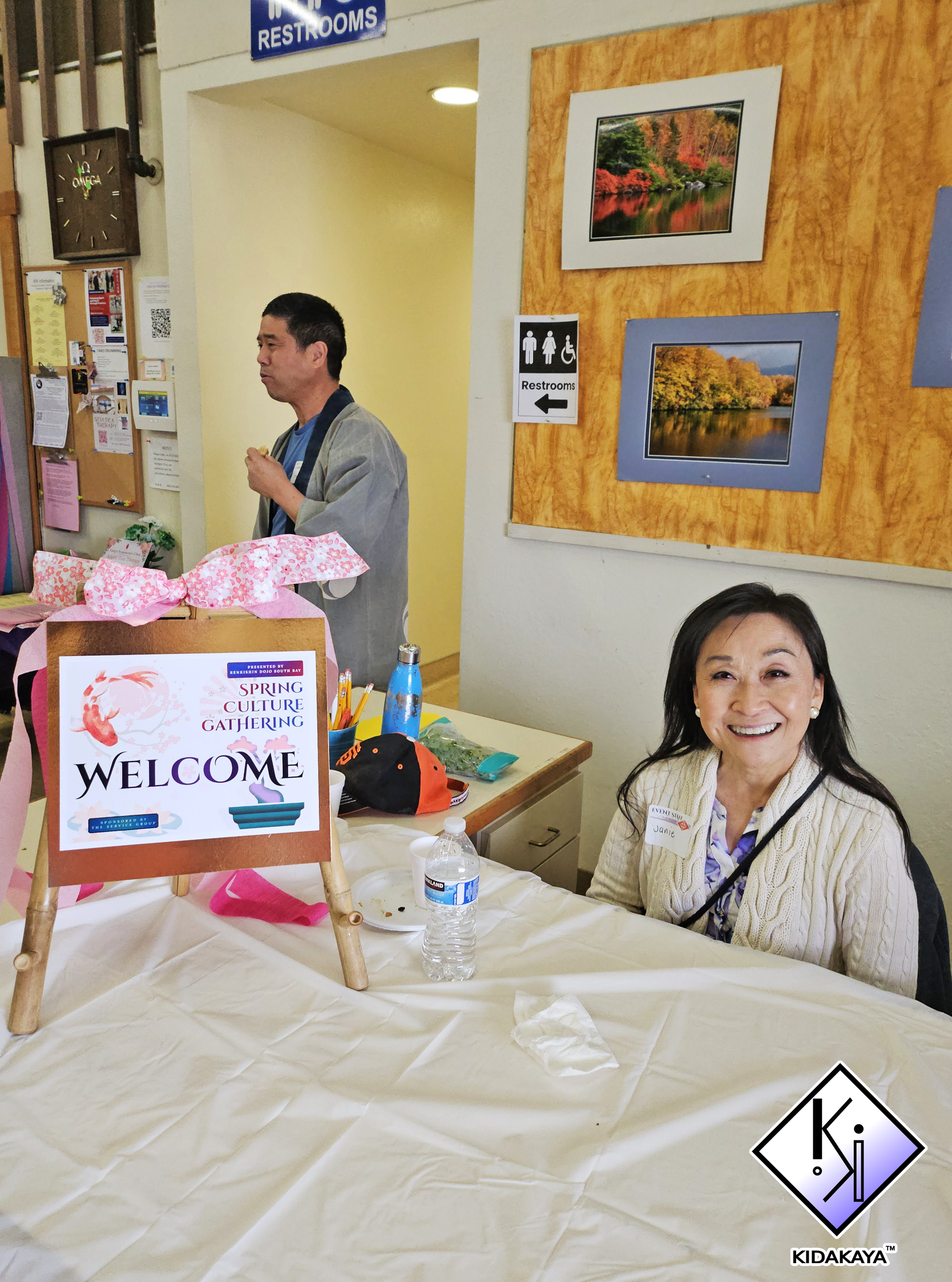 A woman with long black hair and earrings sitting at a table, smiling, in front of a welcome sign for a Spring Culture Gathering event. There is a water bottle, a black cap with orange on it, and miscellaneous items on the table. Behind her, a man wi