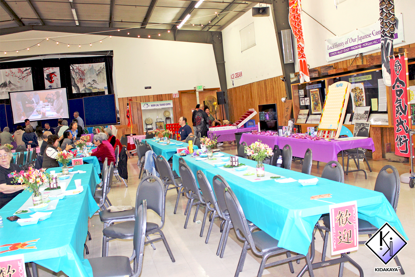 Indoor event with long tables covered in turquoise and purple tablecloths, decorated with flower arrangements, set for a celebration or gathering. People are seated at the tables, some engaged in conversation. A stage with a screen and traditional Ja