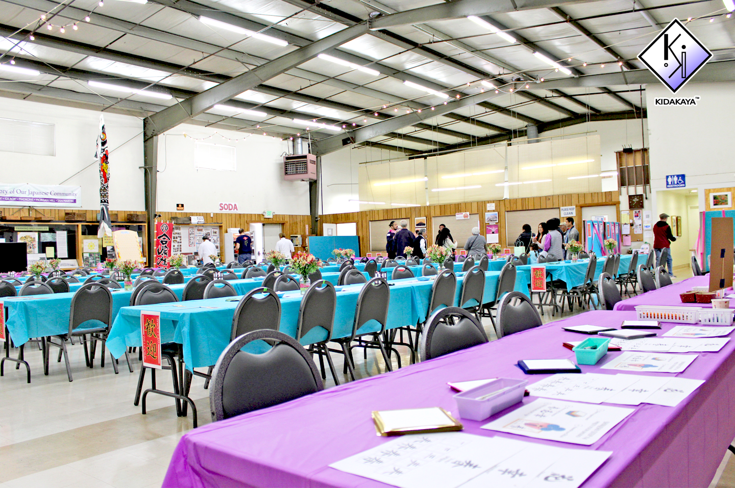 Indoor event space set up with tables covered in blue and purple tablecloths, arranged with flower centerpieces, and surrounded by gray chairs. People are walking and browsing booths along the back wall, which features signs and displays. The room ha