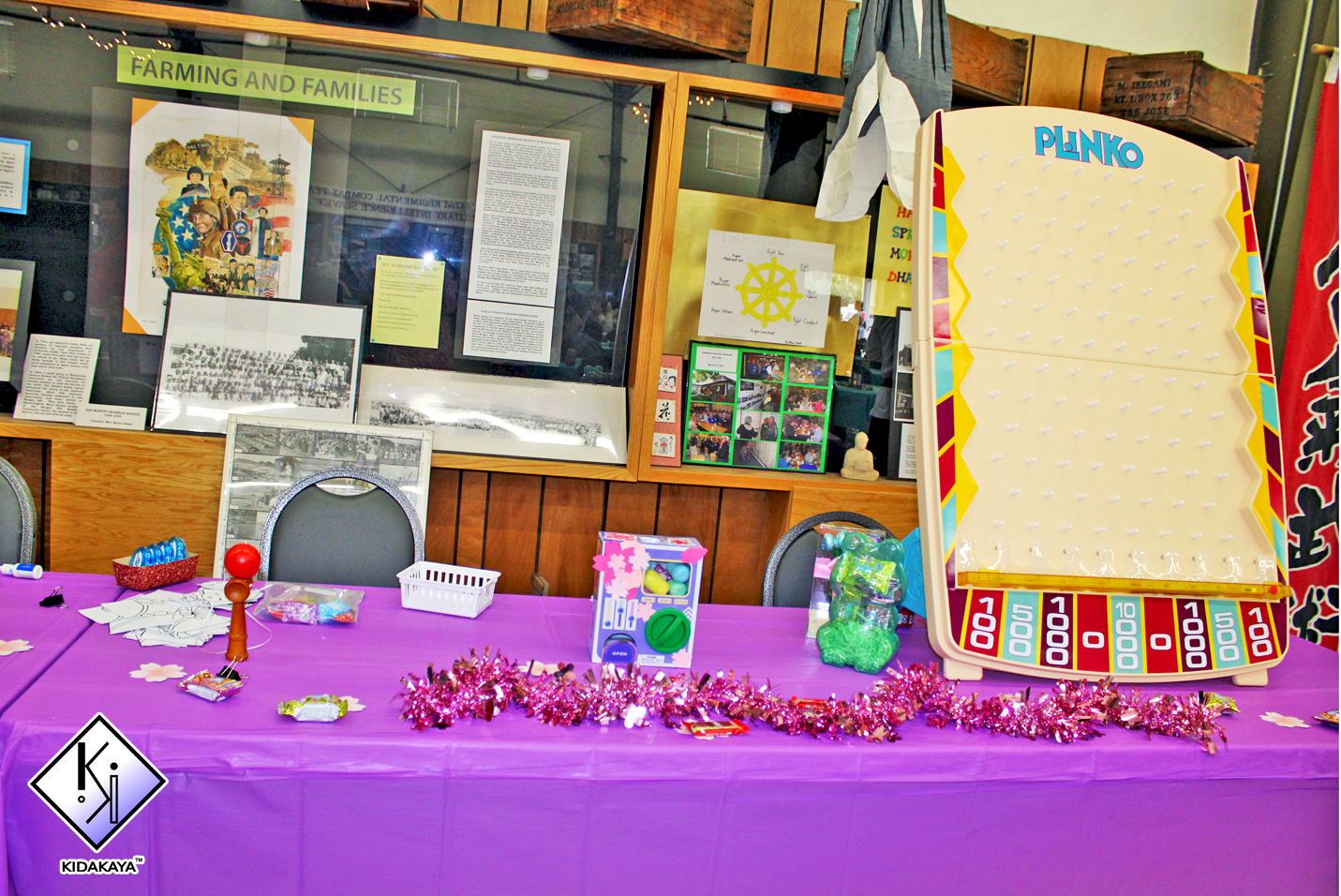 Table decorated with a purple tablecloth, pink tinsel, and small candies, with an arcade game and chairs behind it, inside a room with framed posters and photo displays on the wall, labeled 'Farming and Families'.