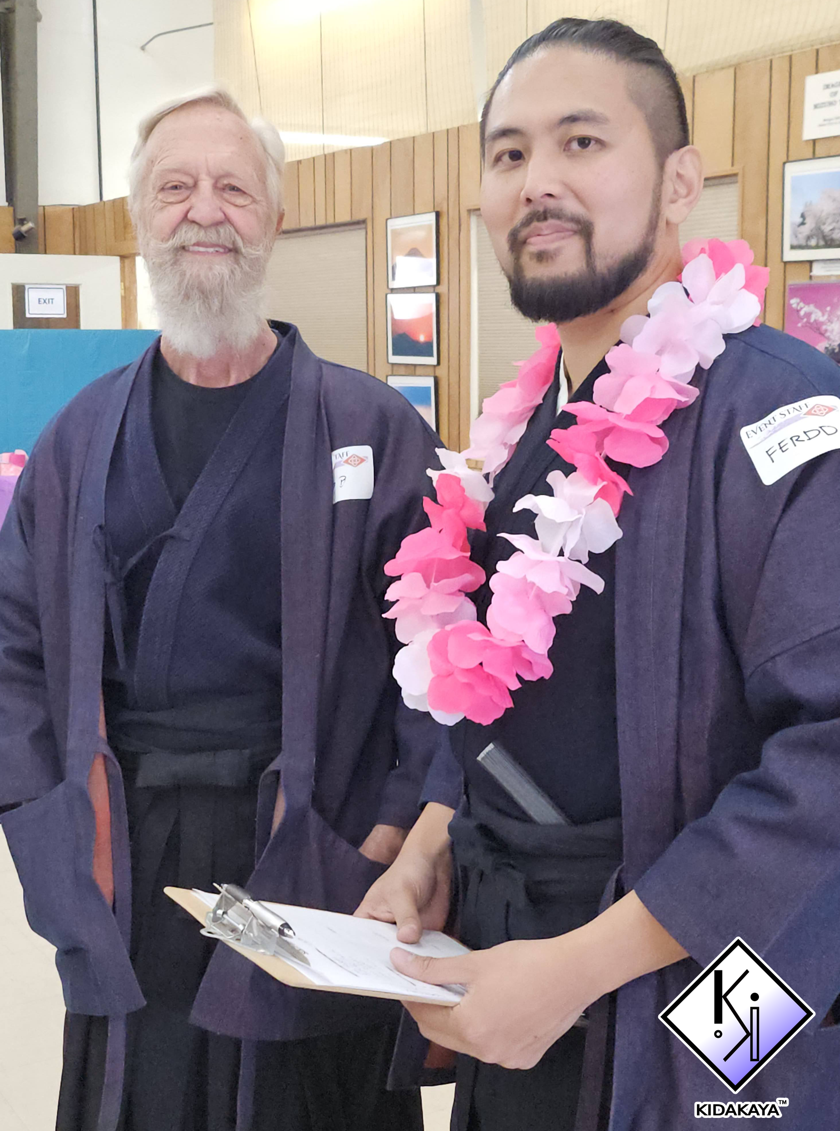 Two men in traditional Japanese attire shaking hands. One man has a white beard and is smiling, while the other has a dark beard and is wearing a pink and white flower lei around his neck, with a serious expression. They are in an indoor setting with