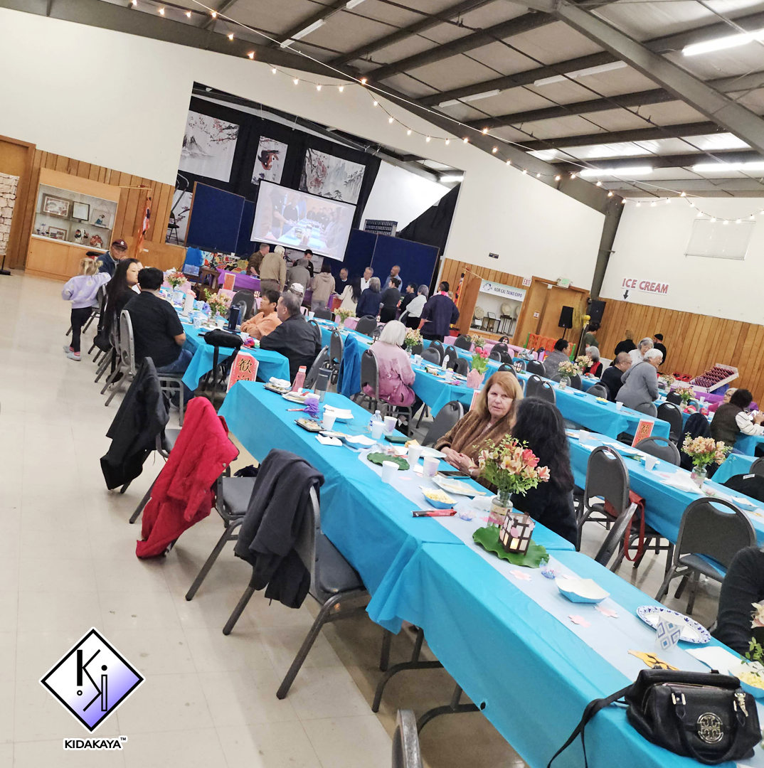 Indoor event with long tables draped in bright blue tablecloths, decorated with flower centerpieces, set for a meal with plates, cups, and food. Crowds of people are seated and mingling, with a stage area at the back featuring a large screen, a podiu