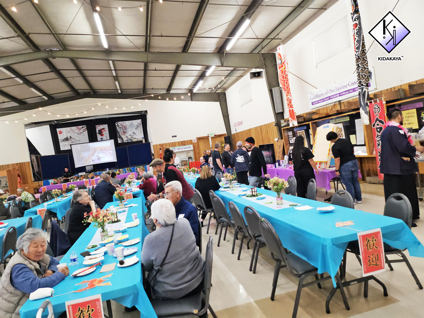Community gathering in a large hall with long tables covered in blue and purple tablecloths, decorated with flowers and featuring food and beverages, with people sitting, standing, and serving, and screens and banners in the background showcasing Jap