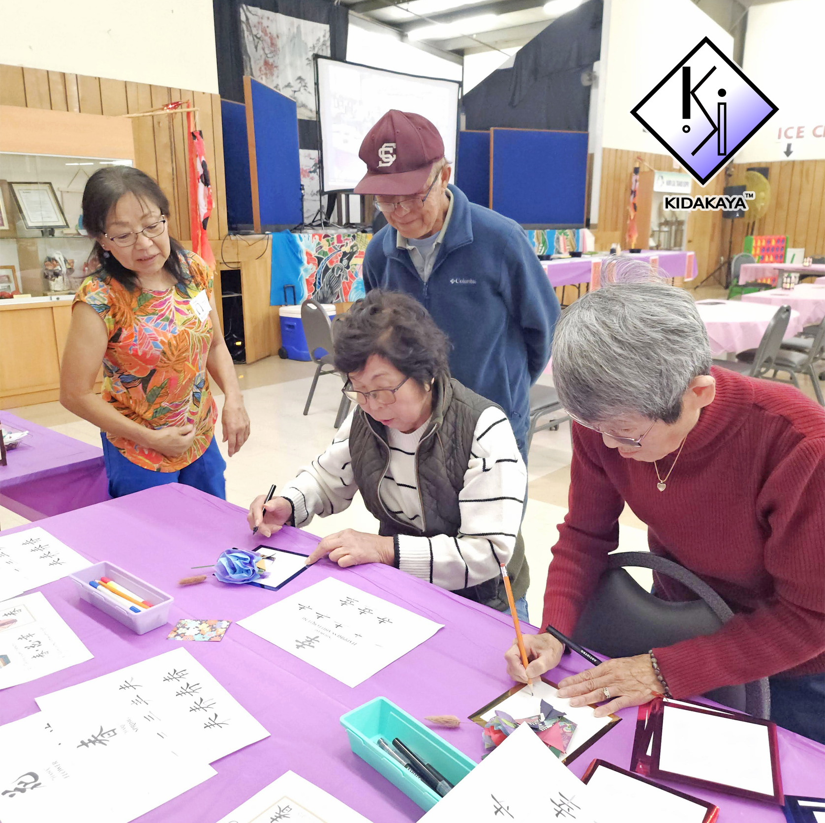 A group of elderly people gathered around a purple table, practicing calligraphy with brushes and black ink, in a community center with decorations and tables in the background.