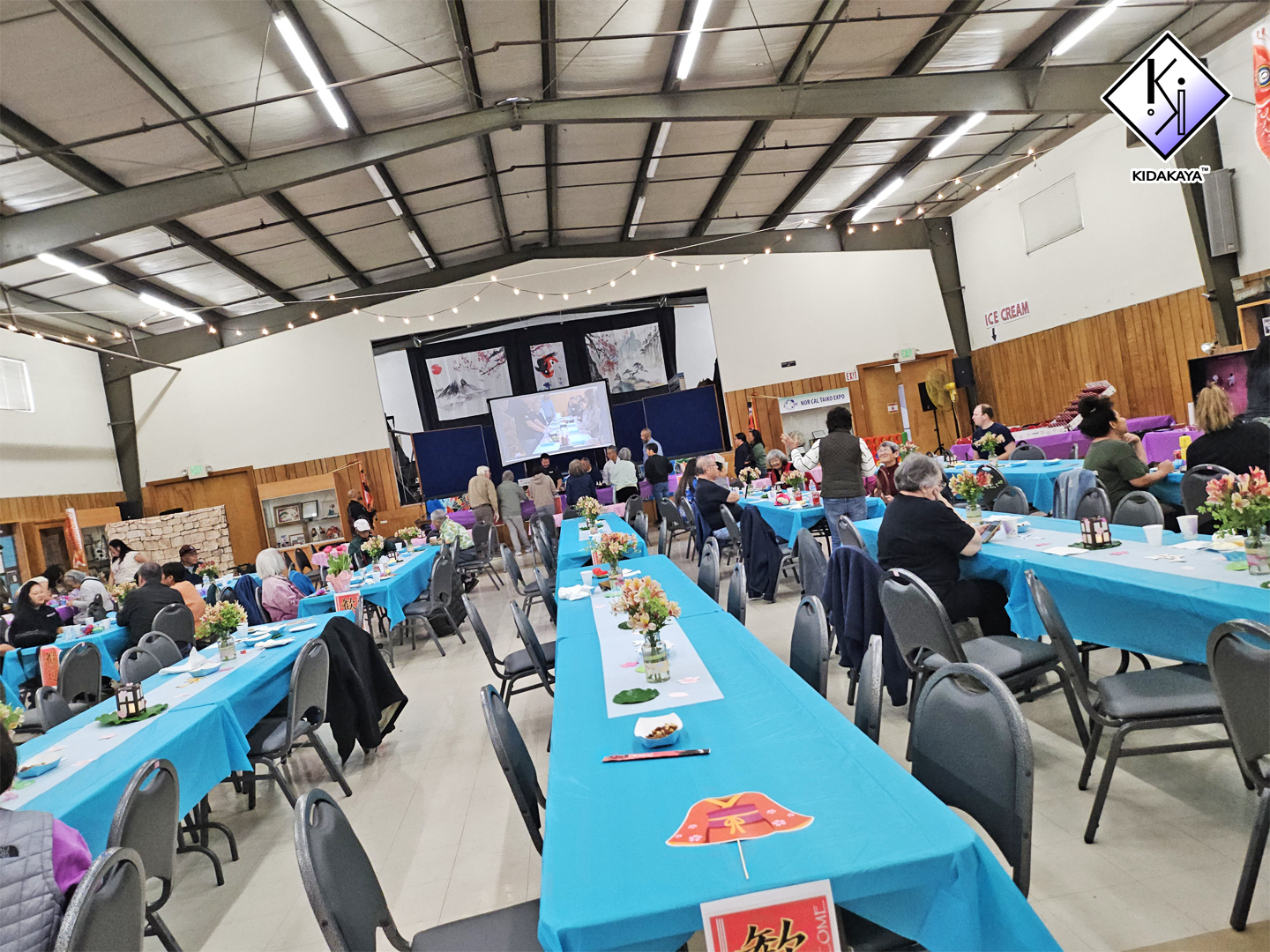 A spacious indoor event hall decorated for a celebration with long tables covered in bright blue tablecloths, floral centerpieces, and paper fans. Attendees are seated and socializing, with some people standing near a stage at the front that has a la