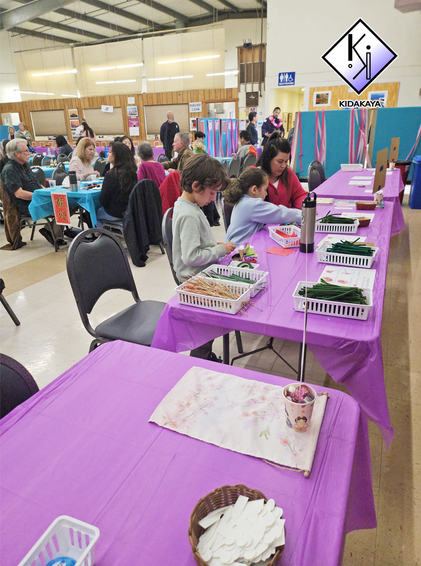Community center indoor event with purple tablecloths, craft supplies like paper and scissors, and diverse people sitting at tables, some working on crafts.