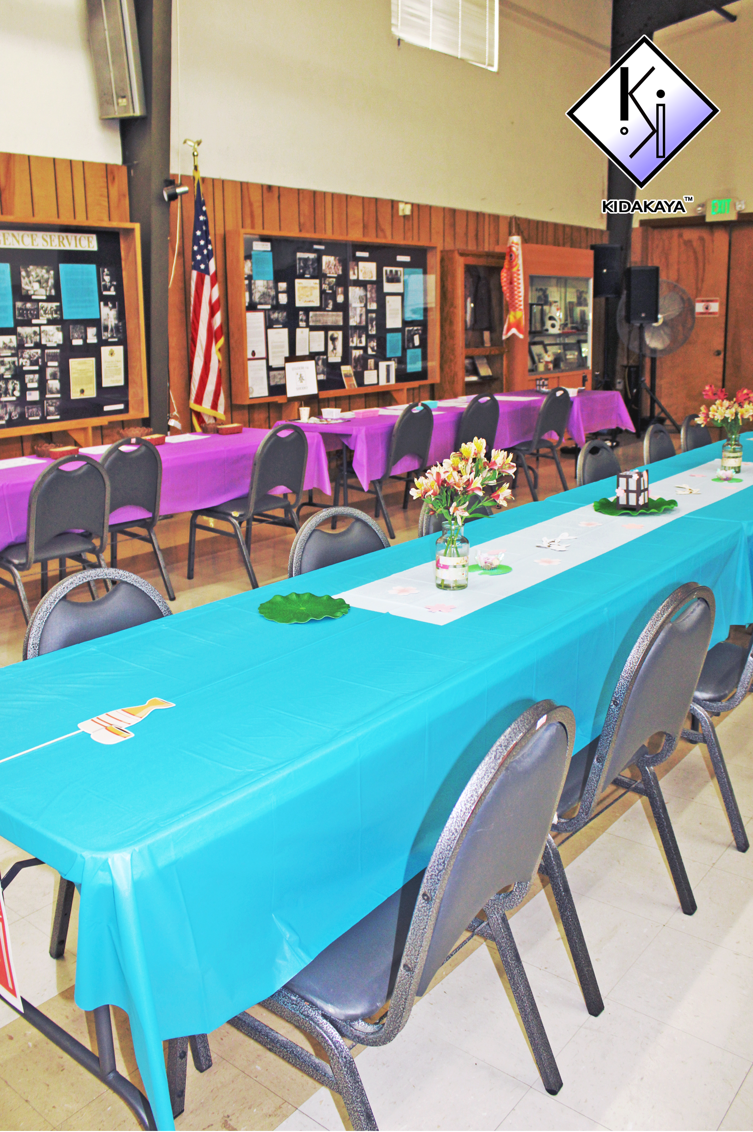 Indoor event space with tables decorated for a celebration, featuring purple and teal tablecloths, floral centerpieces, and assorted decorations on the tables. There are chairs arranged around the tables, a display case, and framed display boards on 
