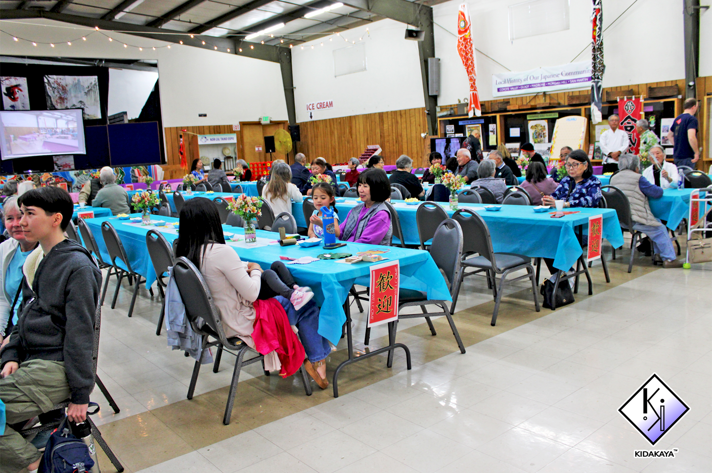 People seated at tables with blue tablecloths inside a hall decorated with Japanese cultural items including koinobori and artwork, some eating, others talking, with a stage and video screens in the background.