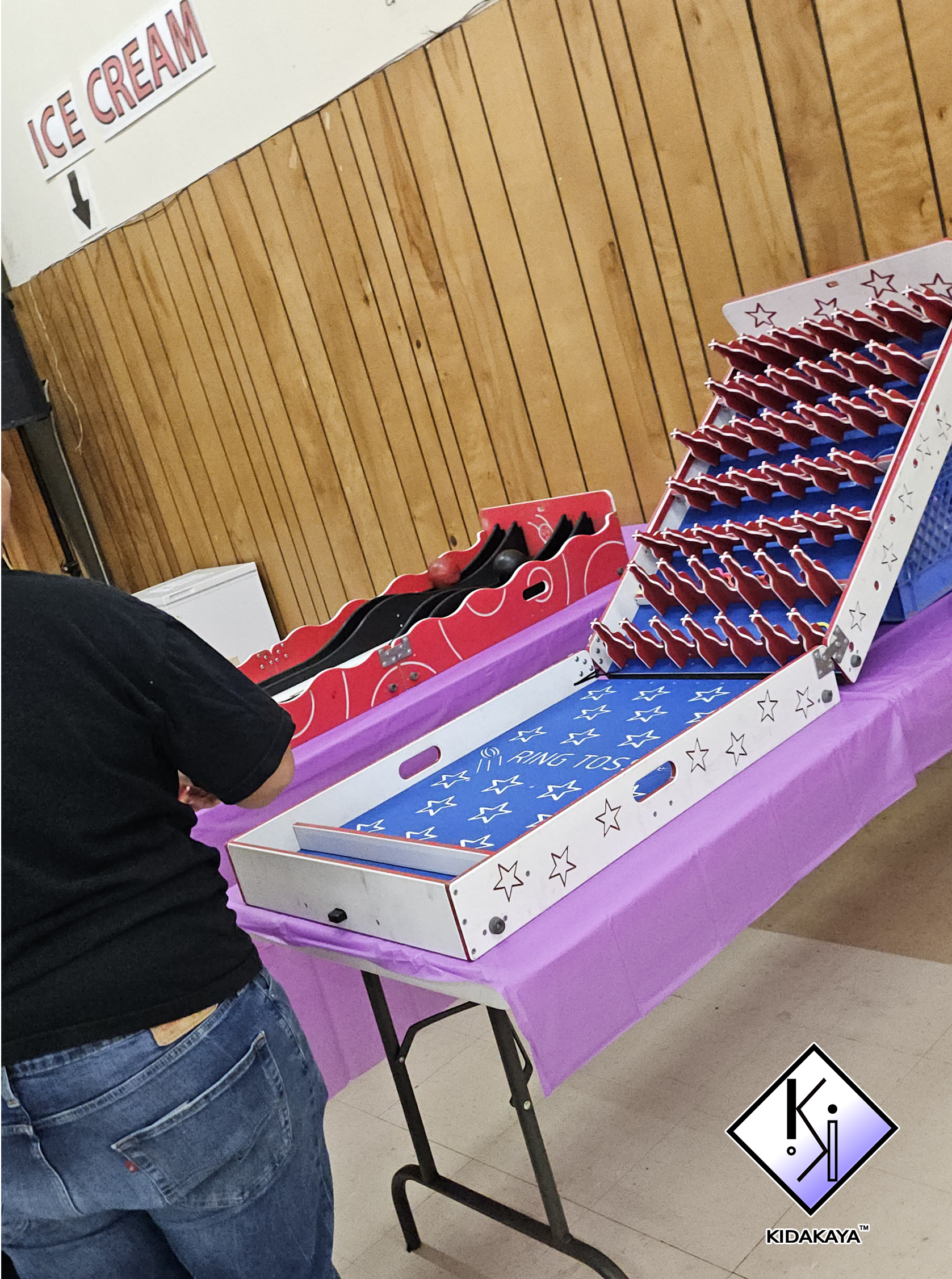 A man in a black shirt and jeans is playing a tabletop game that involves knocking down red and blue wooden sticks with a small black ball, at an event with a purple tablecloth and a wooden-paneled wall in the background.
