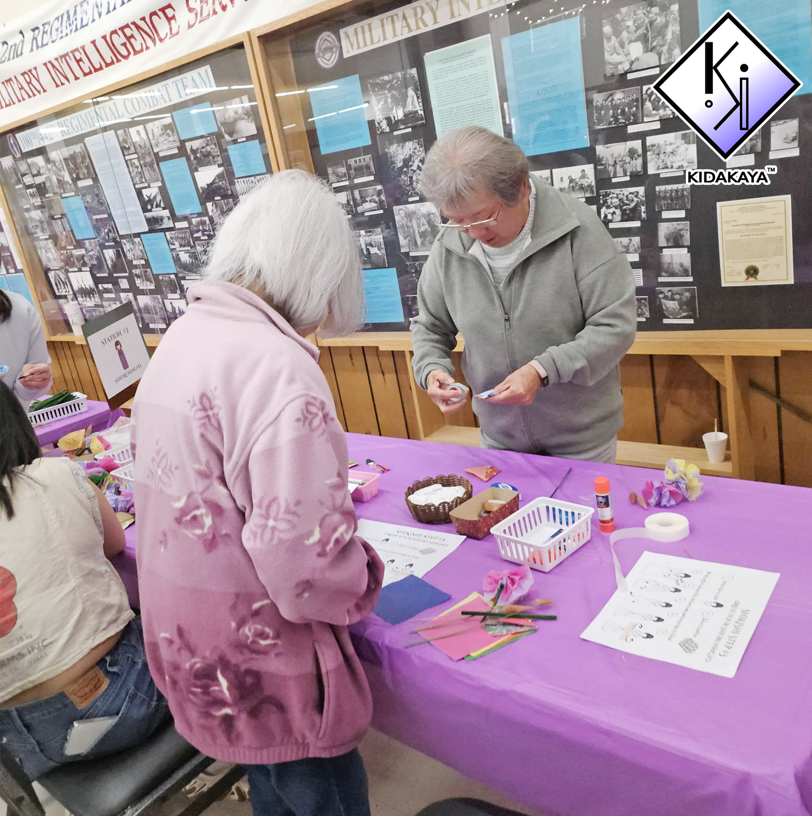 People participating in a craft or arts activity at a table covered with purple tablecloth, with supplies like paper, glue, and flowers, inside a room with display boards about military history in the background.