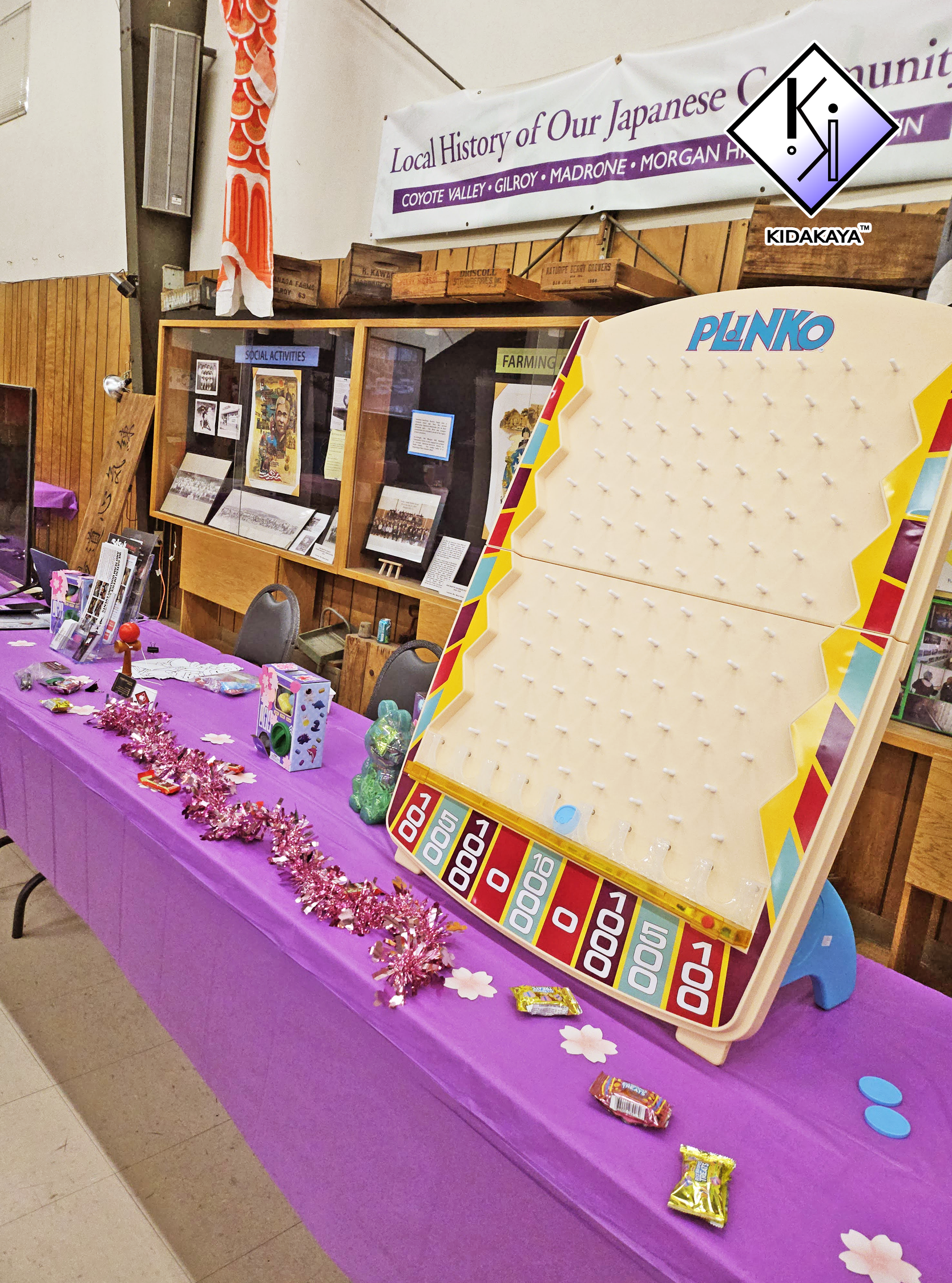 A charity or community event setup with a purple tablecloth decorated with pink tinsel and paper cherry blossom flowers. On the table are small prizes, candies, and a game board with a sliding ball maze. The background features a display case with hi