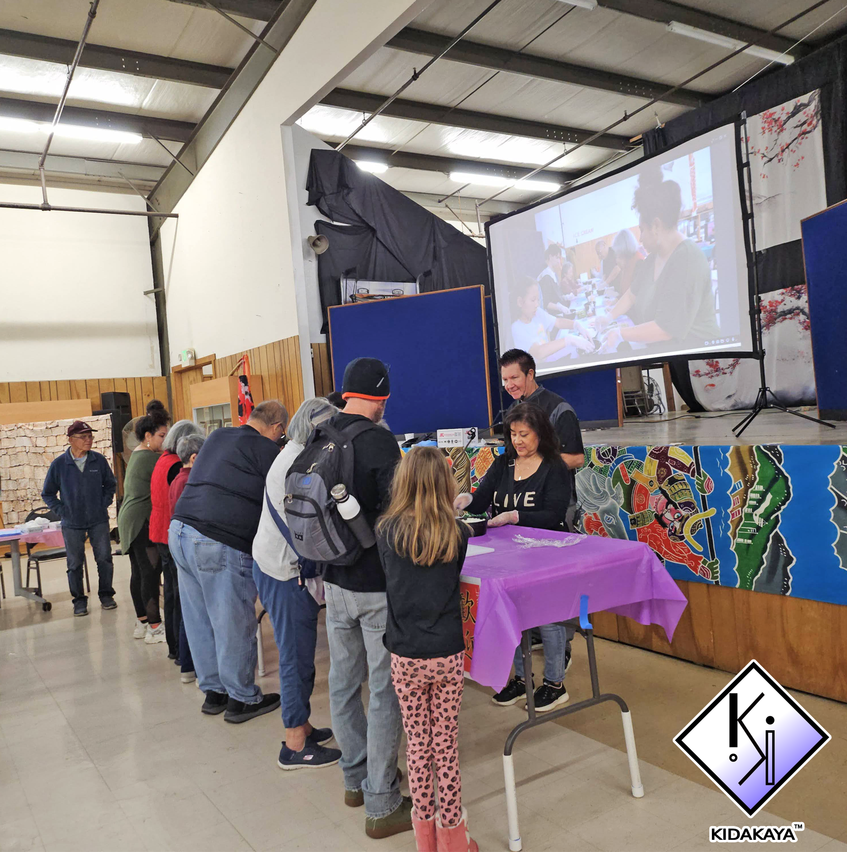 People standing in line at a registration or event table with a purple tablecloth and a colorful decorative banner. A giant screen in the background displays a group of people, including children and adults, engaging in an activity indoors with a hig