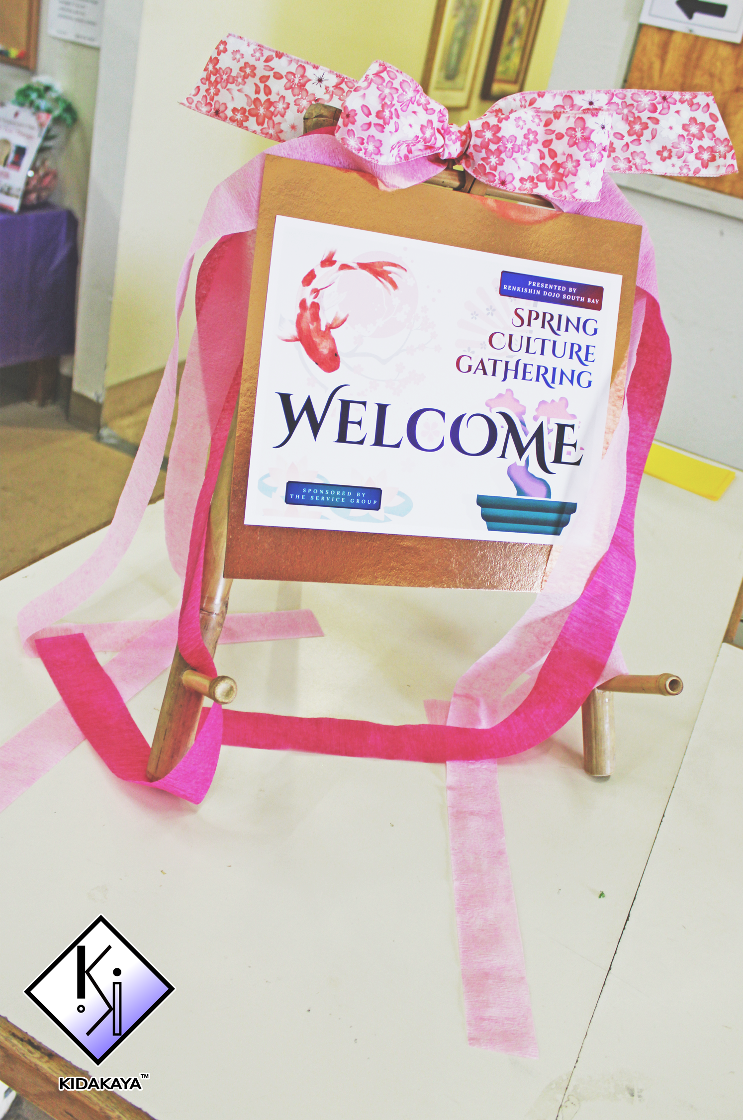 A welcome sign for a Spring Culture Gathering event, attached to a wooden stand with pink ribbons and tissue paper decorations.