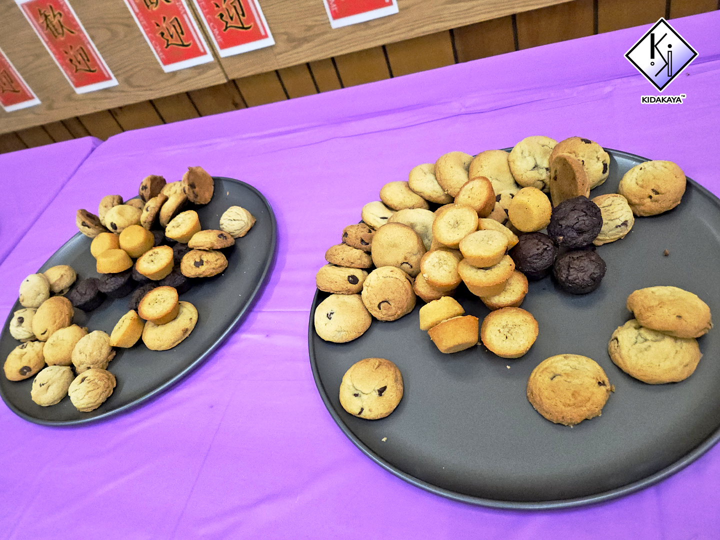 Two black plates with assorted cookies on a purple tablecloth, with red and white signs on a wooden wall in the background.