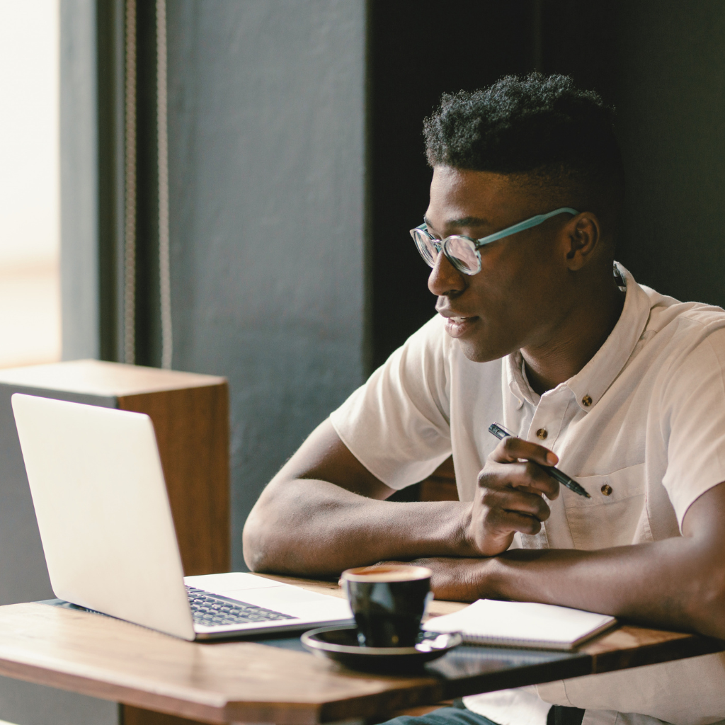 A young man with glasses sitting at a wooden table, using a laptop, holding a pen, with a notebook and a cup of coffee in front of him.