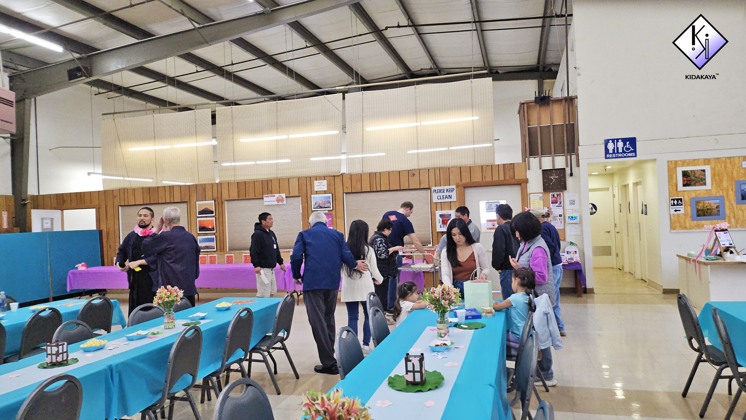 A large indoor event space with people gathered around tables decorated with flowers and treats. There are booths and a sign for restrooms in the background.