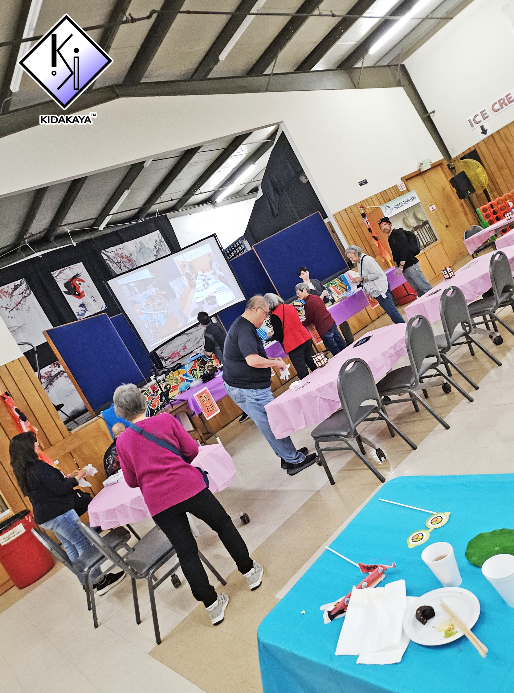 People gathered at a social event with food and decorations, some enjoying sushi. There are tables with pink tablecloths, a blue table with food and chopsticks, and a large screen displaying images. The background has Japanese-themed artwork and a si