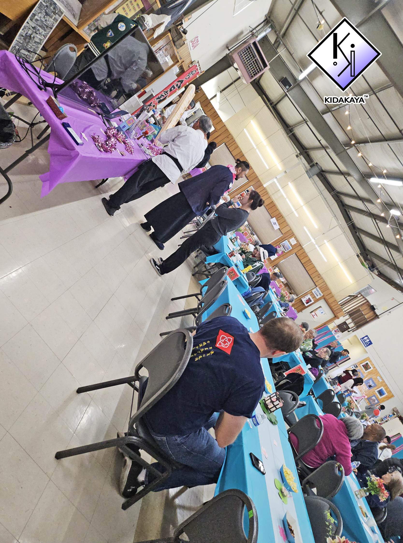 People gathered at tables with purple and blue tablecloths at an indoor event or market, with various displays and merchandise.
