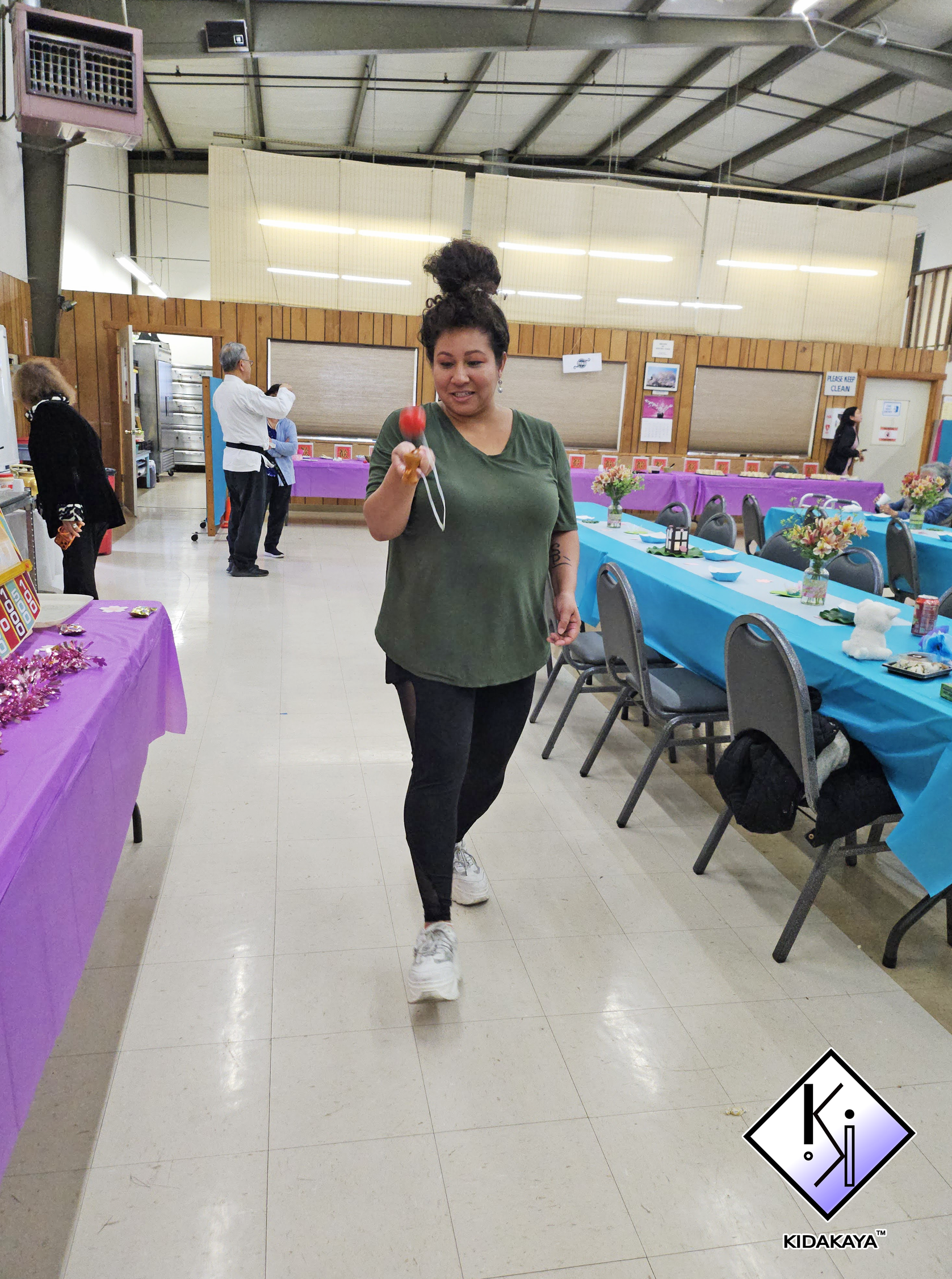 A woman with a high messy bun hairstyle is walking in a decorated indoor event space, holding a red and orange spinning top toy. The room has tables with purple and turquoise tablecloths, decorated with flower arrangements, and people are seen in the