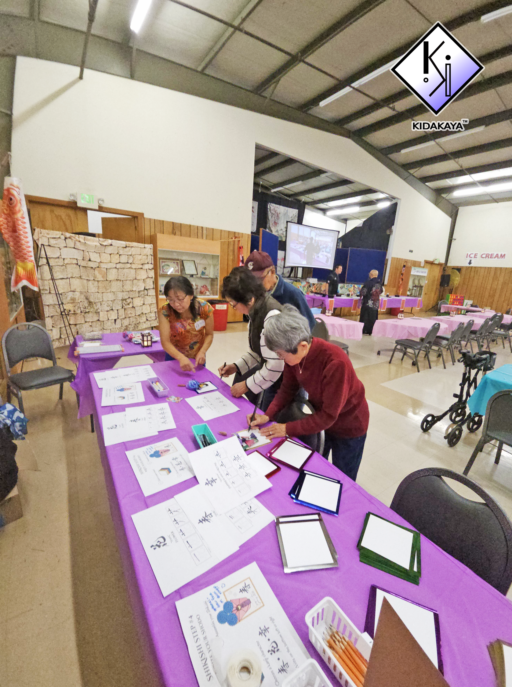 People engaging in a calligraphy workshop at a table covered with purple cloth, inside a community center or hall with beige walls and a high ceiling. Various calligraphy practice sheets and writing supplies are on the table.