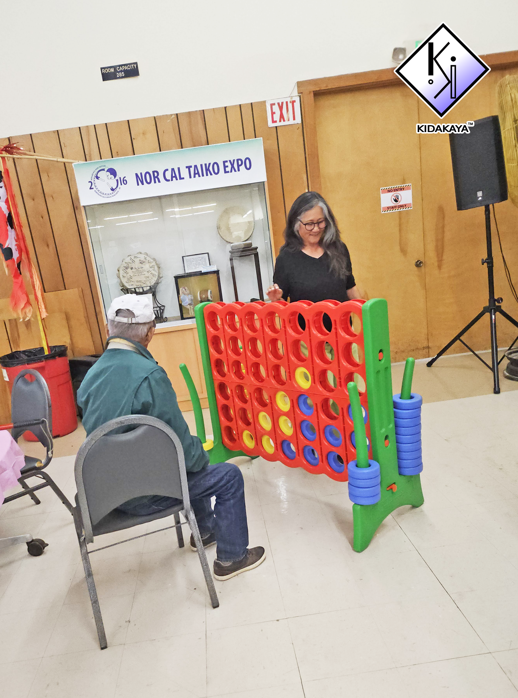 An indoor scene with a person playing a large connect four game, while another person with gray hair and glasses sits on a chair nearby. There are chairs, a trash bin, and a sign for a taiko expo in the background.