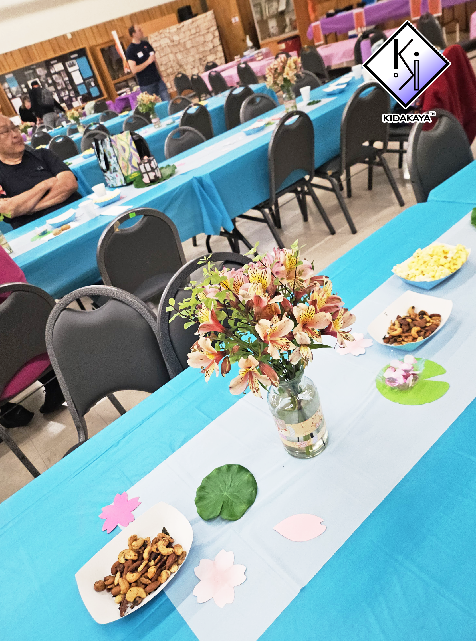 A decorated party table with blue and white tablecloths, pink and green paper cutouts, a vase of pink flowers, and bowls of snacks, set in a hall with other decorated tables and people in the background.