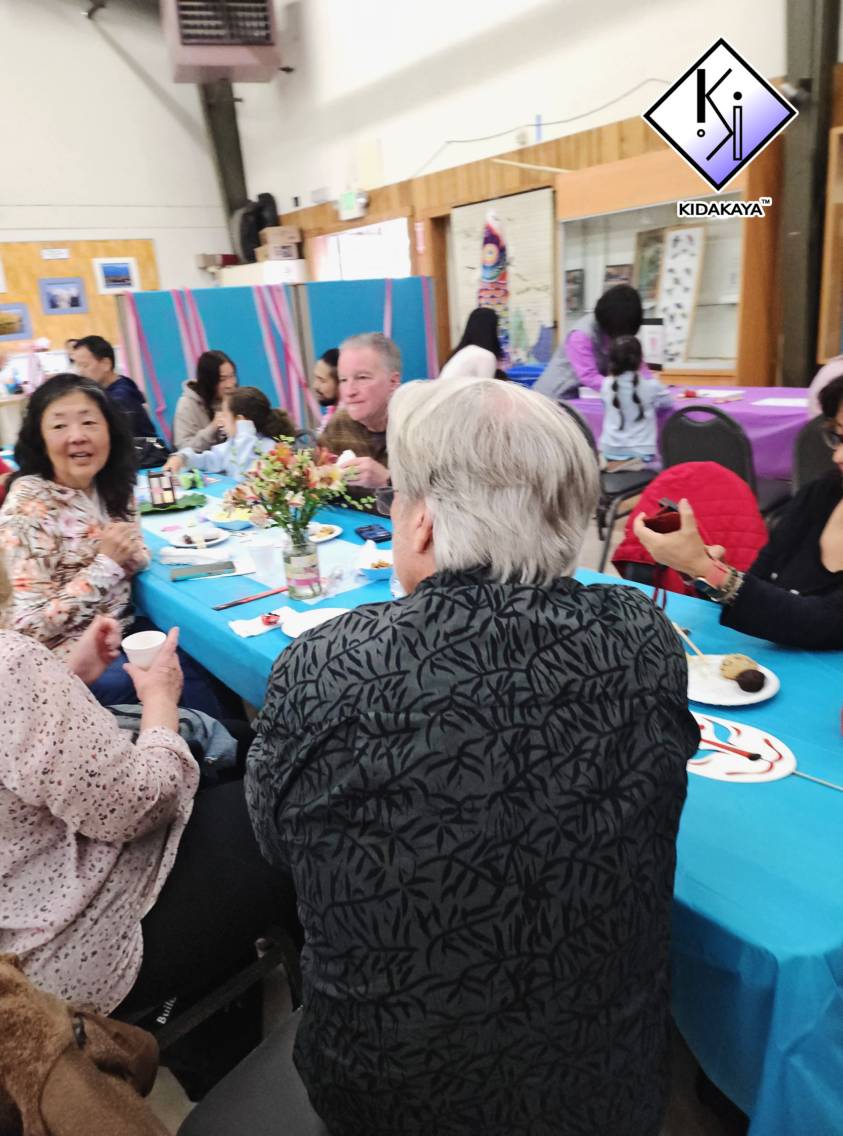 People gathered around a table at a festive celebration, with decorations, food, and drinks, in a room with wooden walls.