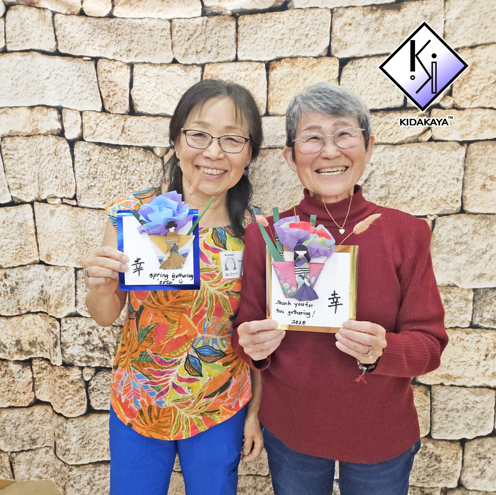 Two women smiling and holding colorful origami flower cards that say 'Thank you for this gathering! 2020' and have Japanese characters on them, standing in front of a stone wall.