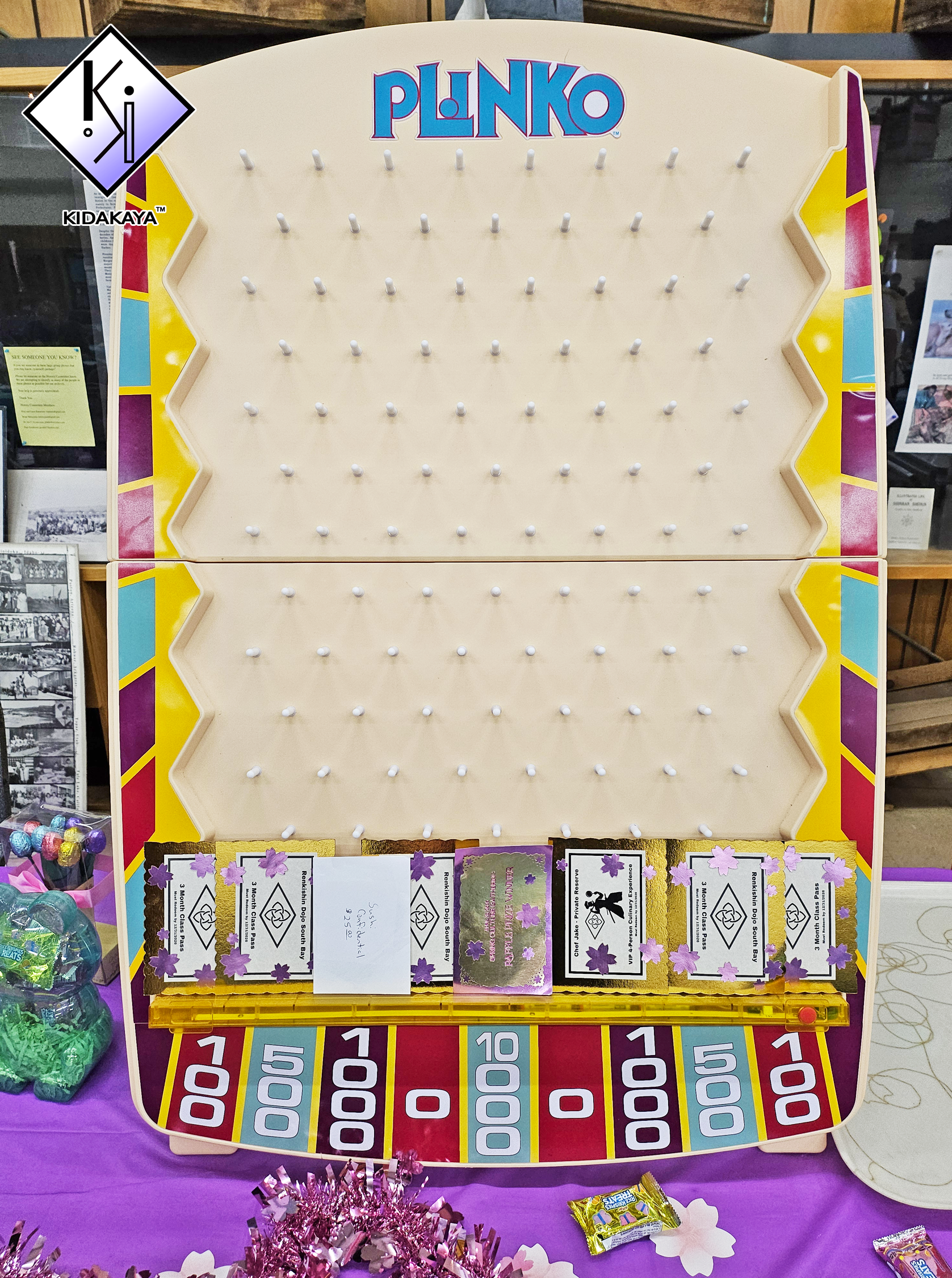 Plinko game board with colorful sections and pegs, decorated with purple and pink crafts, candy, and cards, set on a purple table at a fair or event.
