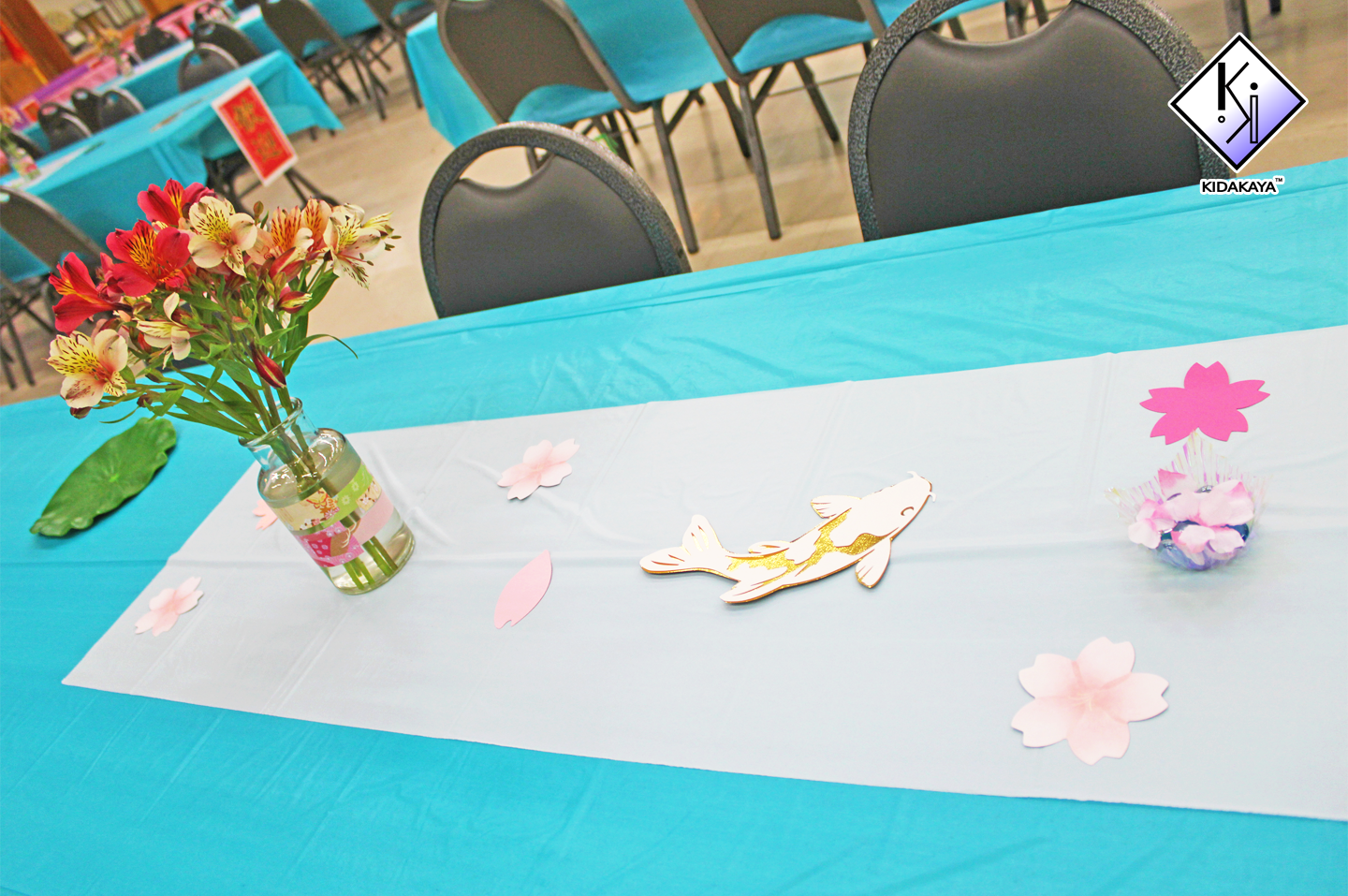 Table decorated with a blue tablecloth, white table runner, pink paper flowers, a fish cutout, and a vase of pink and yellow flowers at a celebration or event.